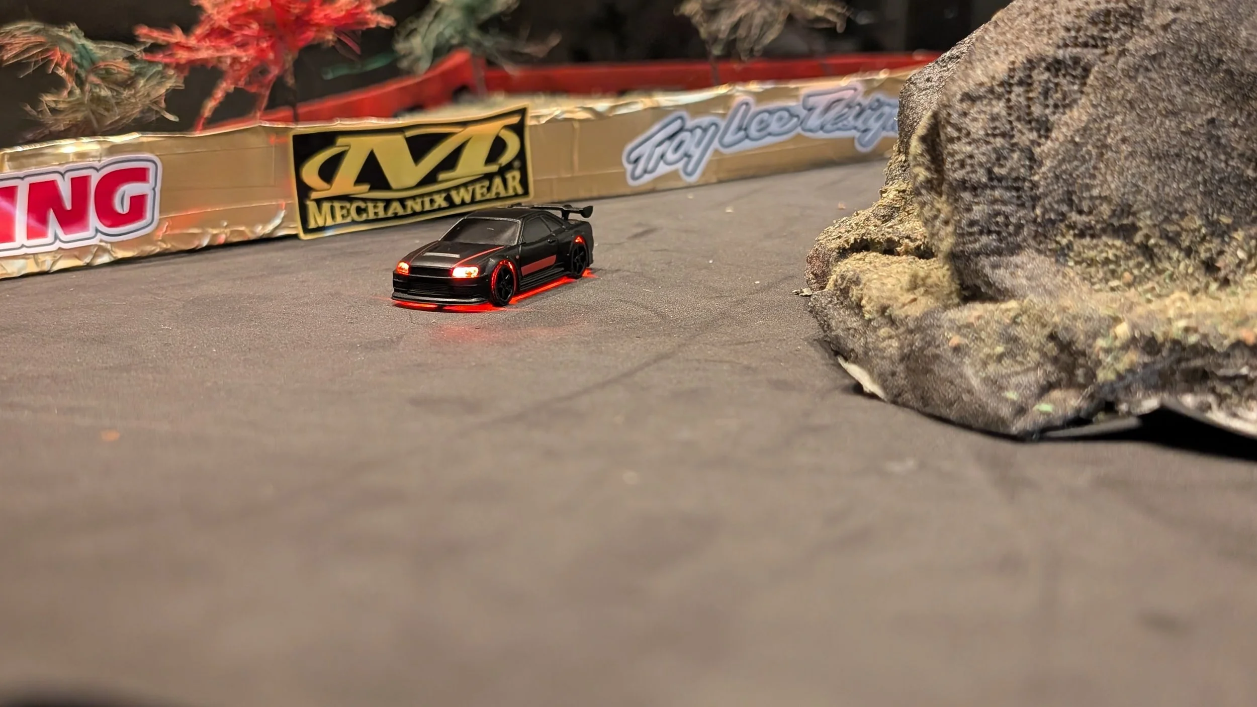 Close-up of a miniature black sports car model racing around the track near a rock, with a background of a rocky terrain and a race track sponsor wall.