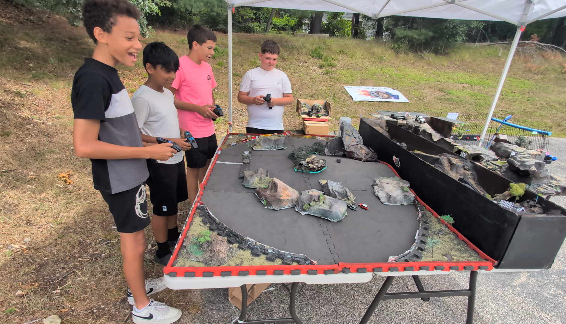 Group of teens having fun racing on a tabletop RC race track outdoors under a canopy, with model rocks, terrain.