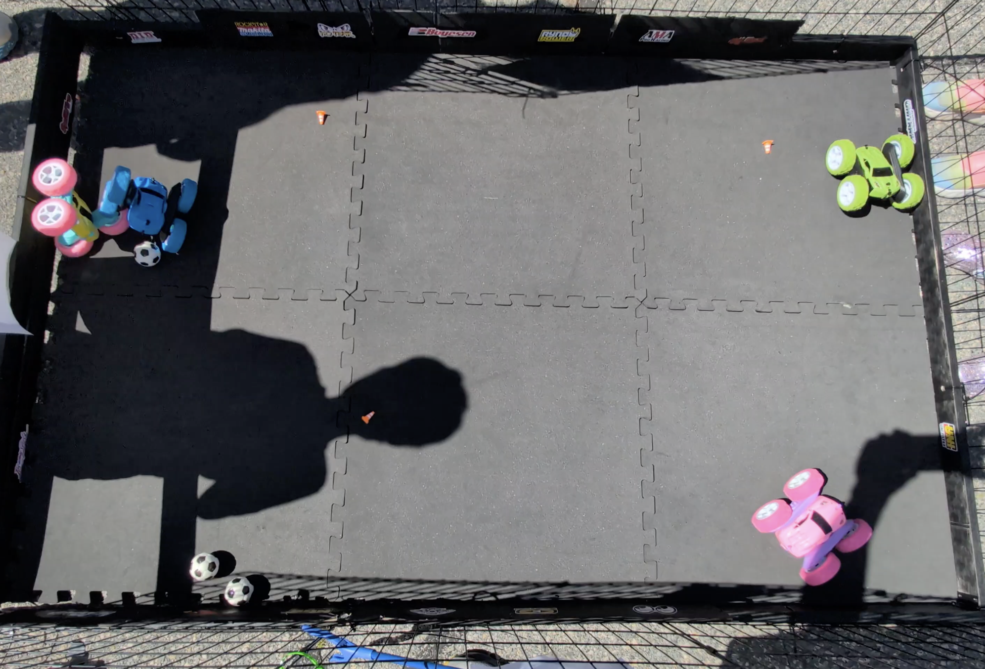 A fenced outdoor play area with artificial grass flooring, containing small toy cars, soccer balls, and small orange cones, with shadows cast on the ground.