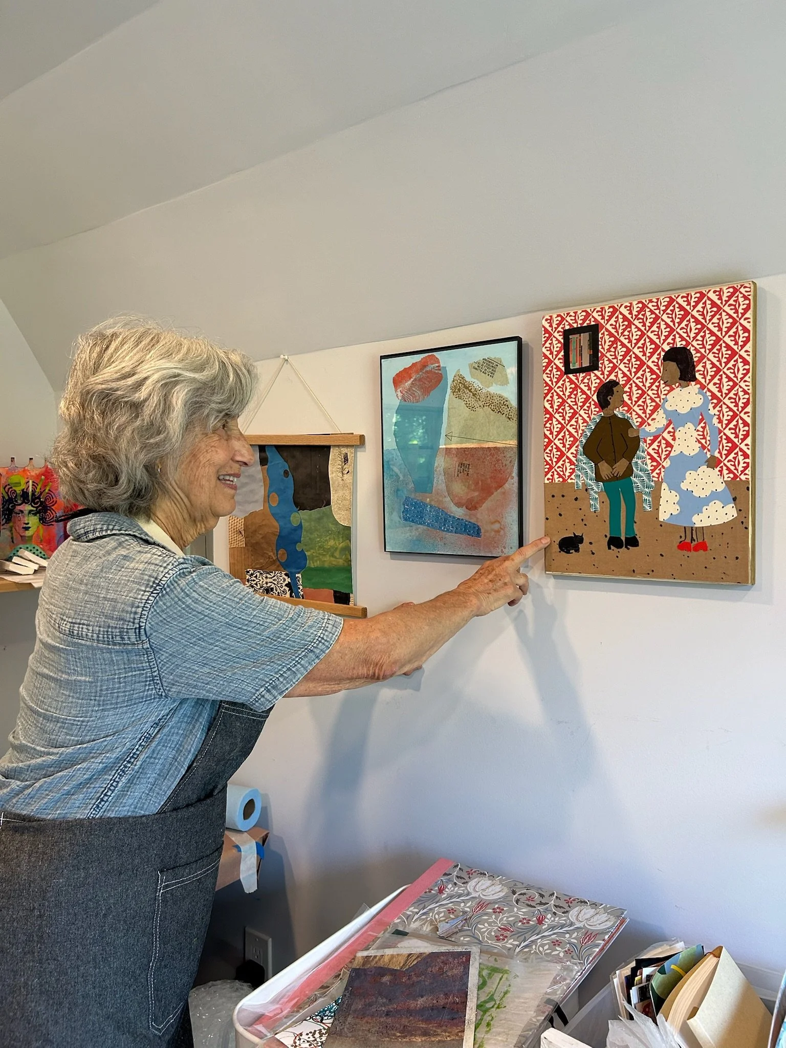 Alice Goodman smiling and pointing at wall art in an art gallery or studio, with colorful paintings and art supplies on a table below.