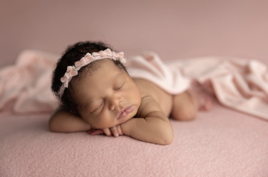 A sleeping newborn baby girl with a pink floral headband, lying on a pink textured blanket with a pink cloth draped over her back.