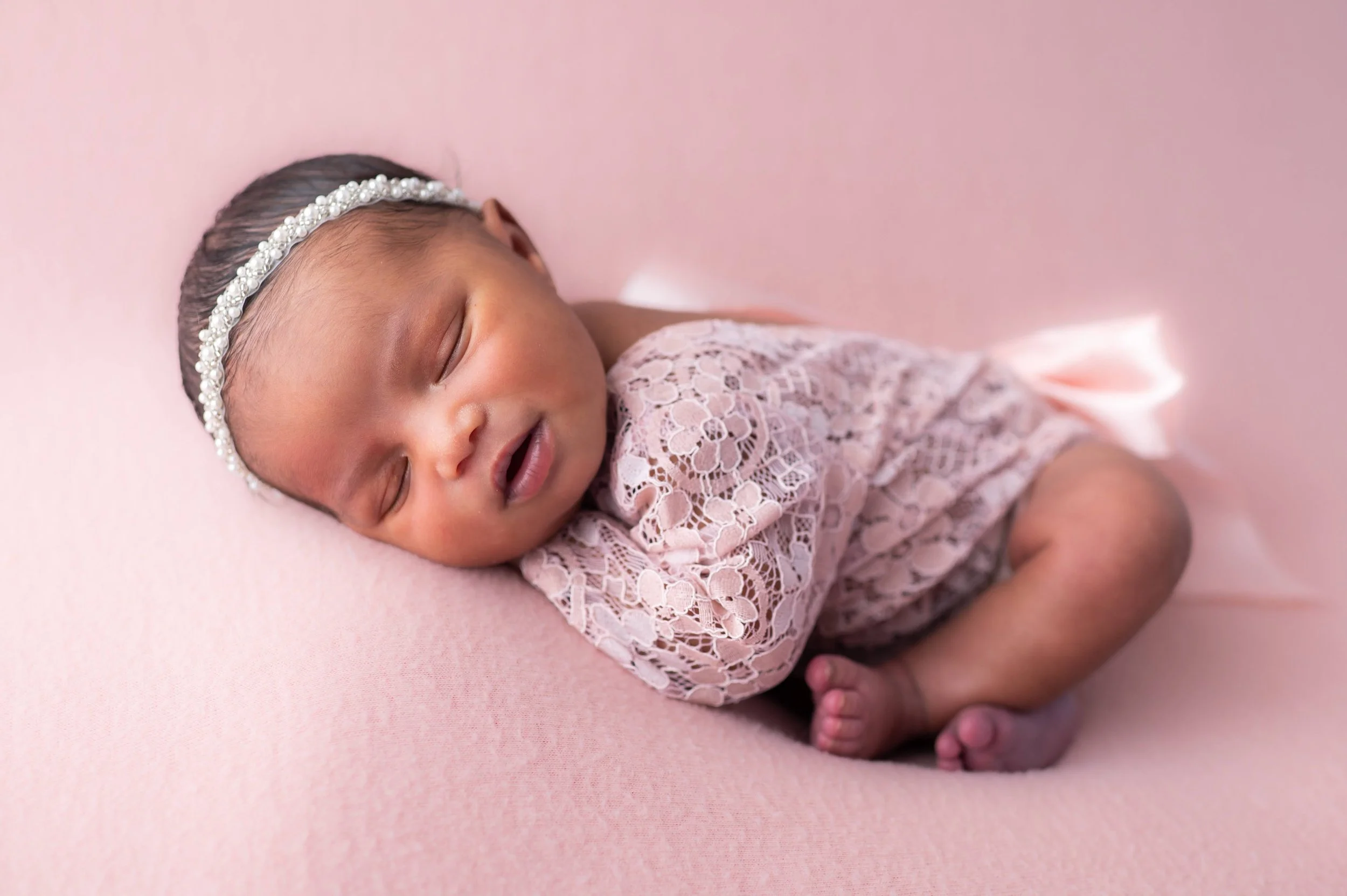 A sleeping baby girl with a pearl headband, wearing a lace pink dress, lying on a soft pink surface.