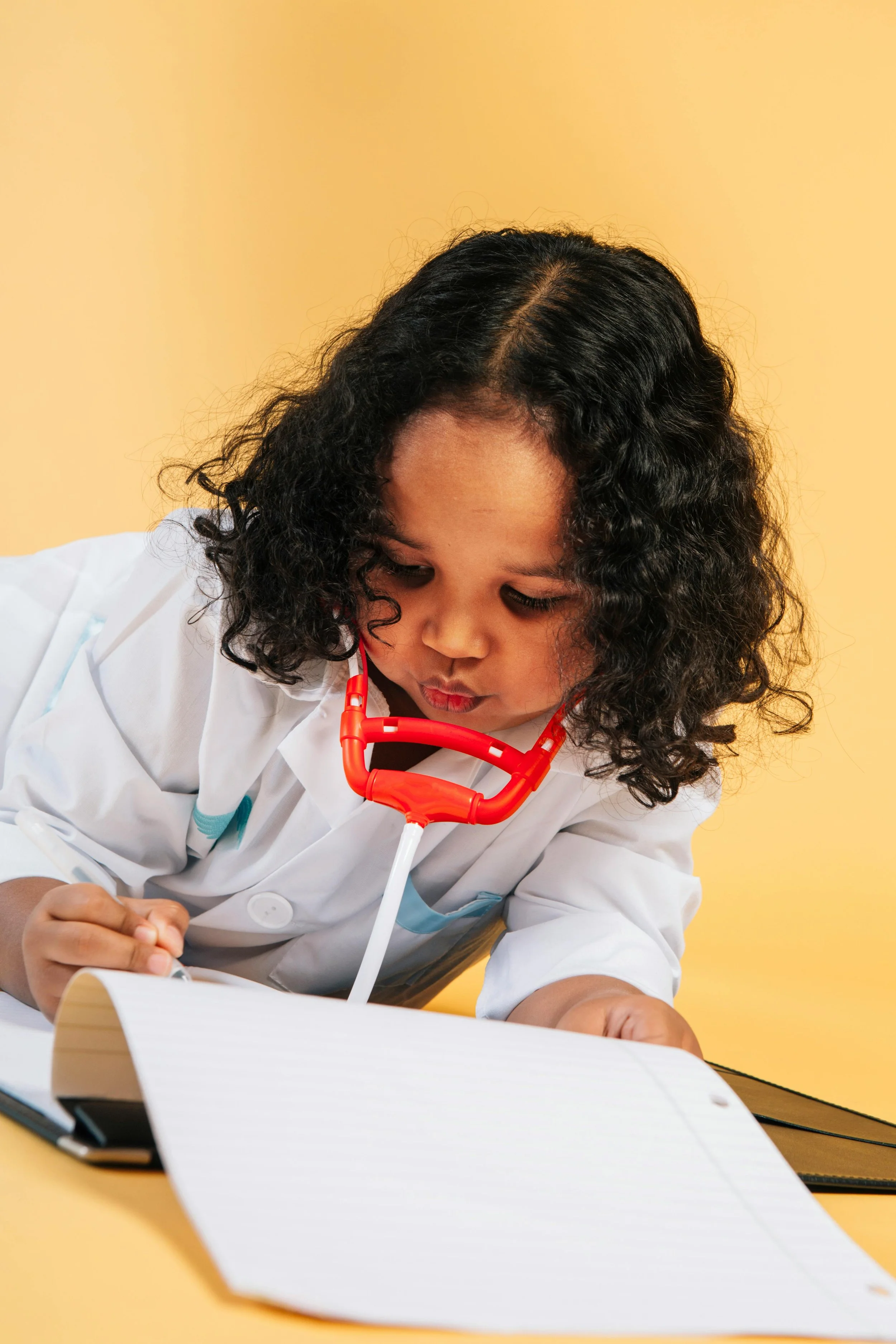 Young girl wearing a white coat and a red stethoscope studying at a table with notebook and pen, against a yellow wall background.