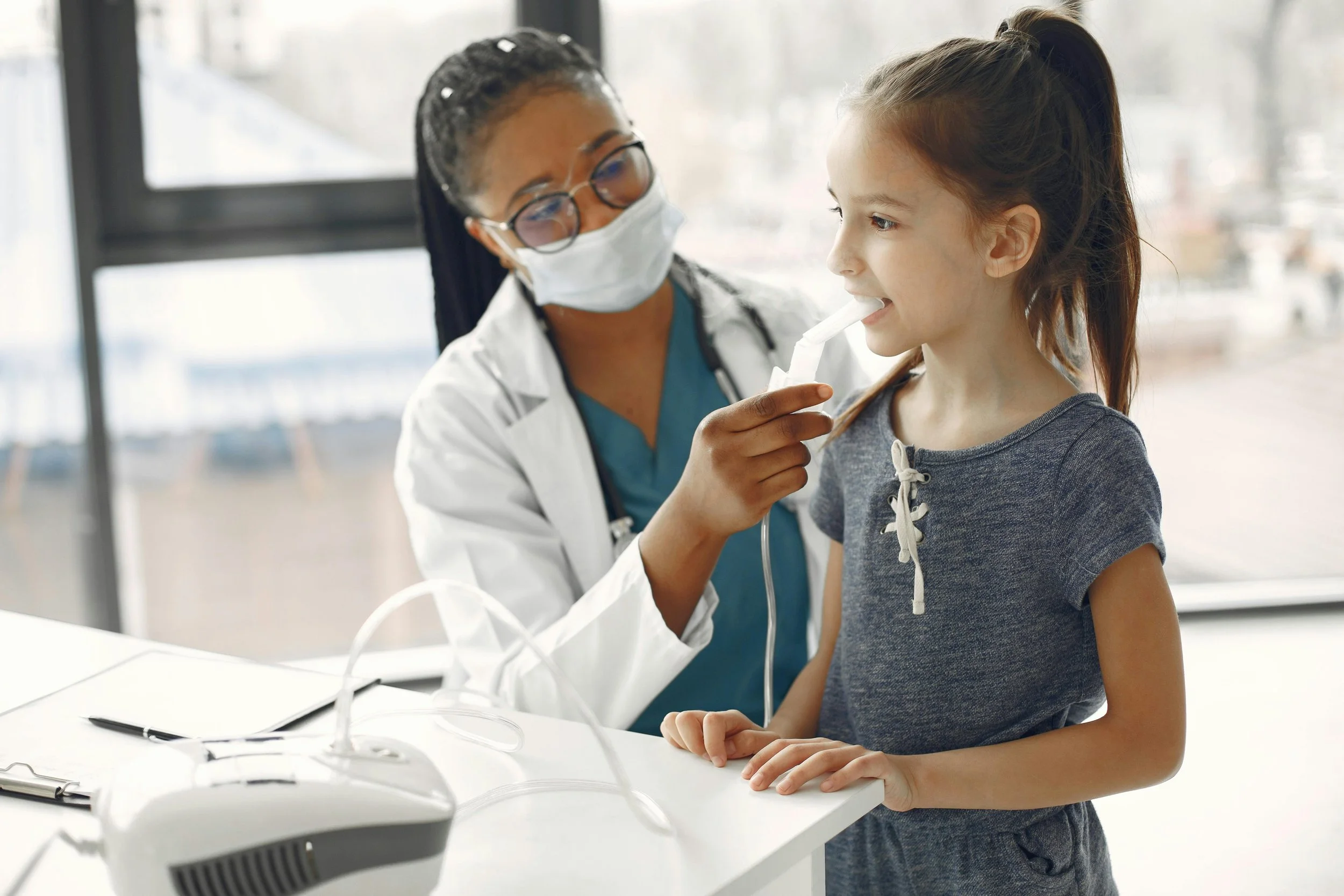 A healthcare worker wearing glasses and a mask is examining a young girl with a stethoscope in a medical office.