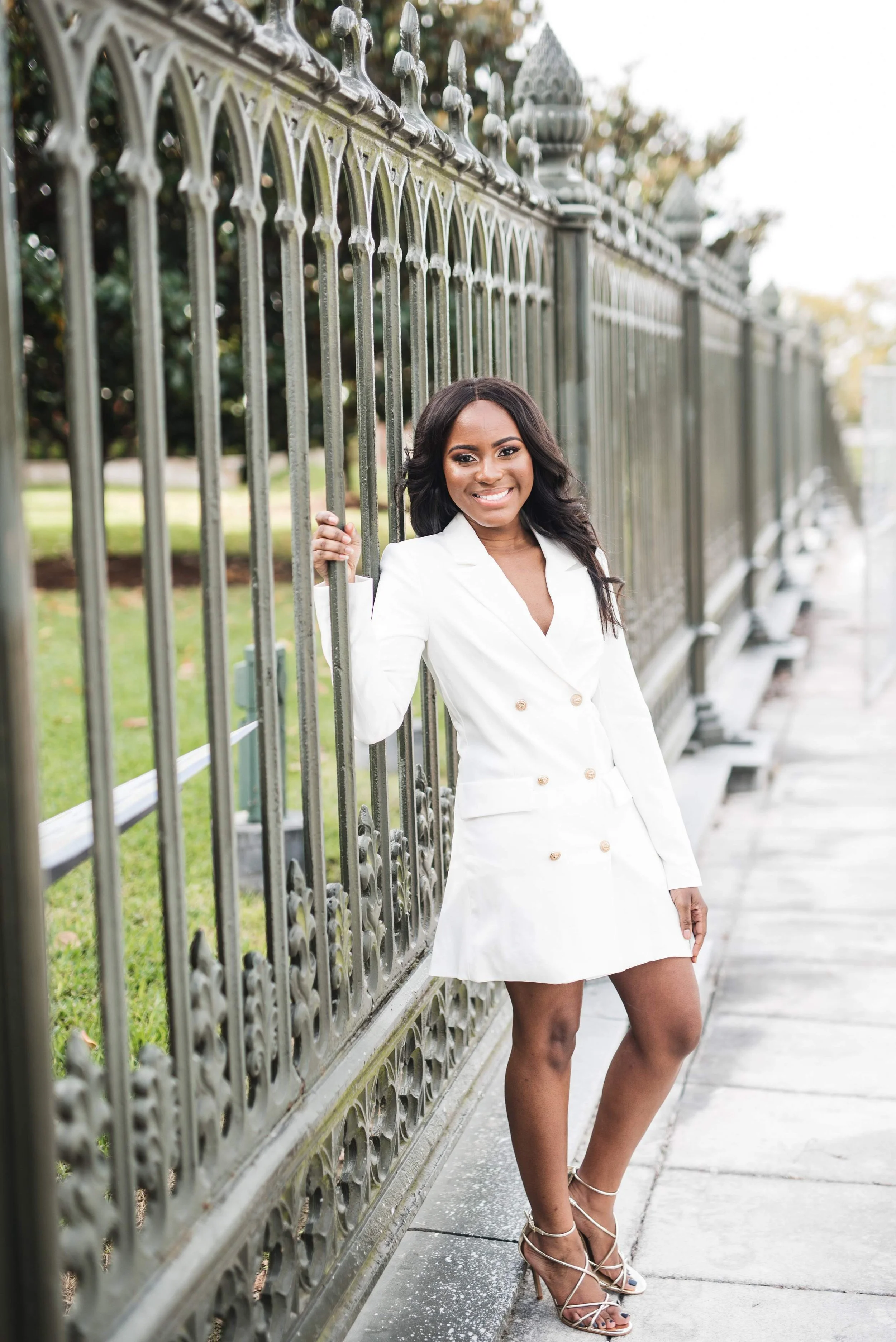 A woman in a white blazer dress and high heels standing next to an ornate metal fence, smiling at the camera.
