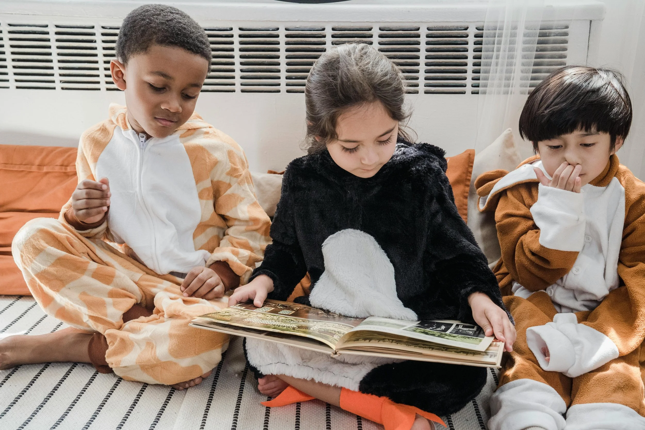 Three children sitting on a striped rug on the floor, looking at a book. They are wearing animal-themed pajamas and are sitting closely together.