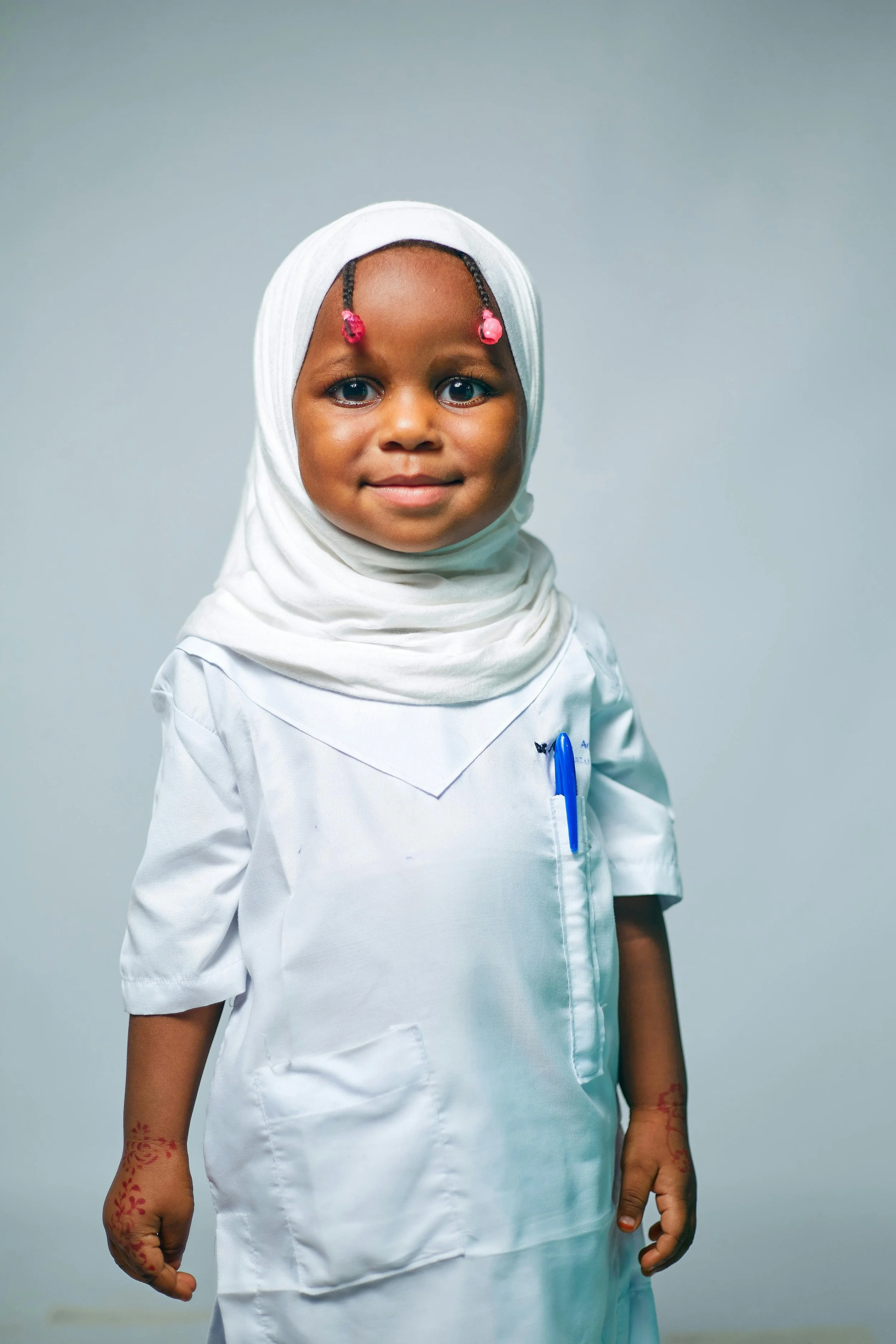 A young girl with braided hair wearing a white hijab and a white medical coat, standing against a plain background.