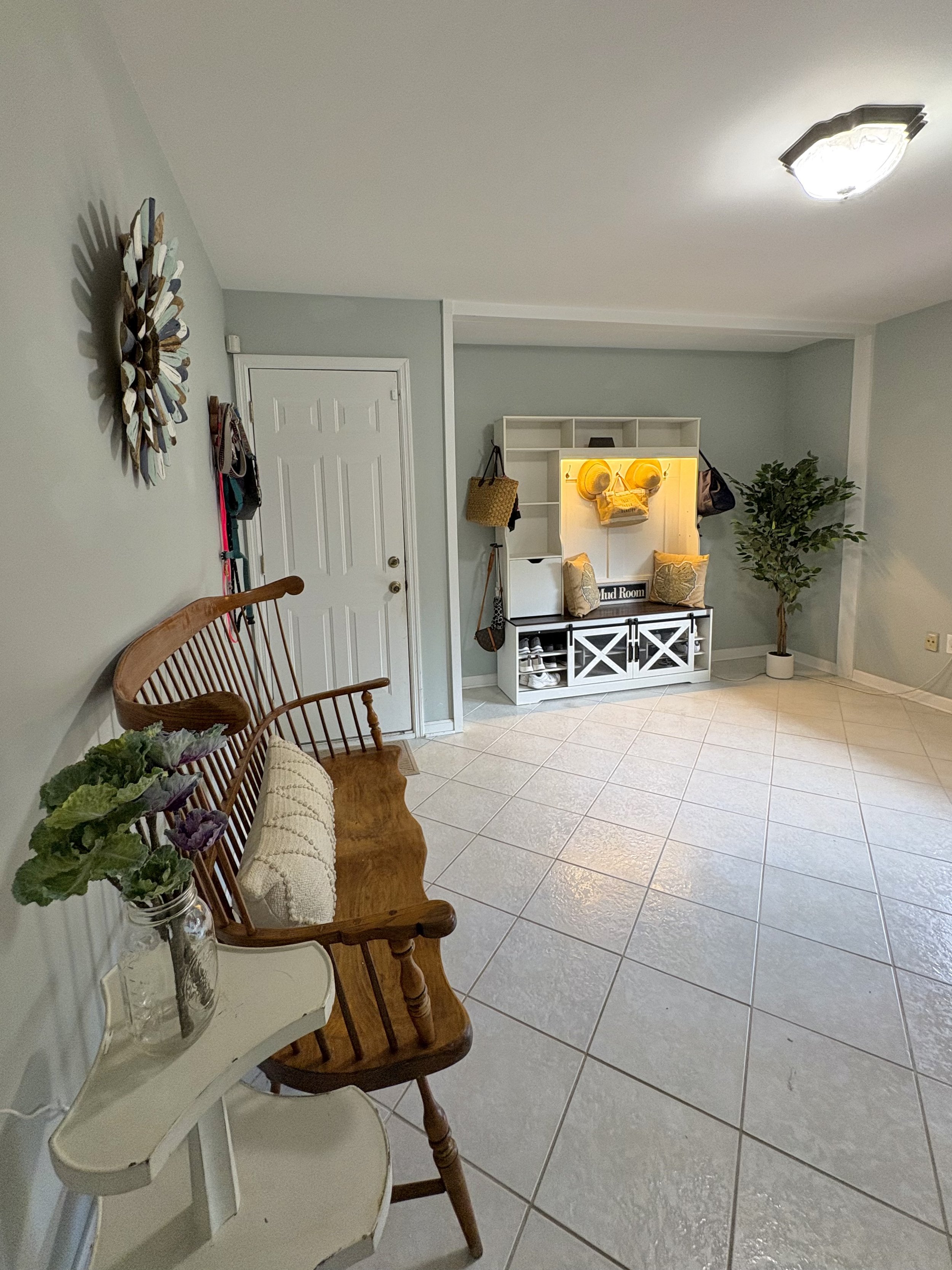 Living room with a white wall, a wooden bench with a beige pillow, a small white table with a glass jar of purple and green flowers, a coat rack with hats and bags, a white shelf with yellow lamps, a large potted plant, and a tiled floor.