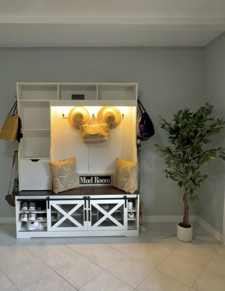 Indoor mudroom with white storage bench, two pillows, a sign reading 'Mud Room', wall hooks with bags, and a tall potted plant on the right.
