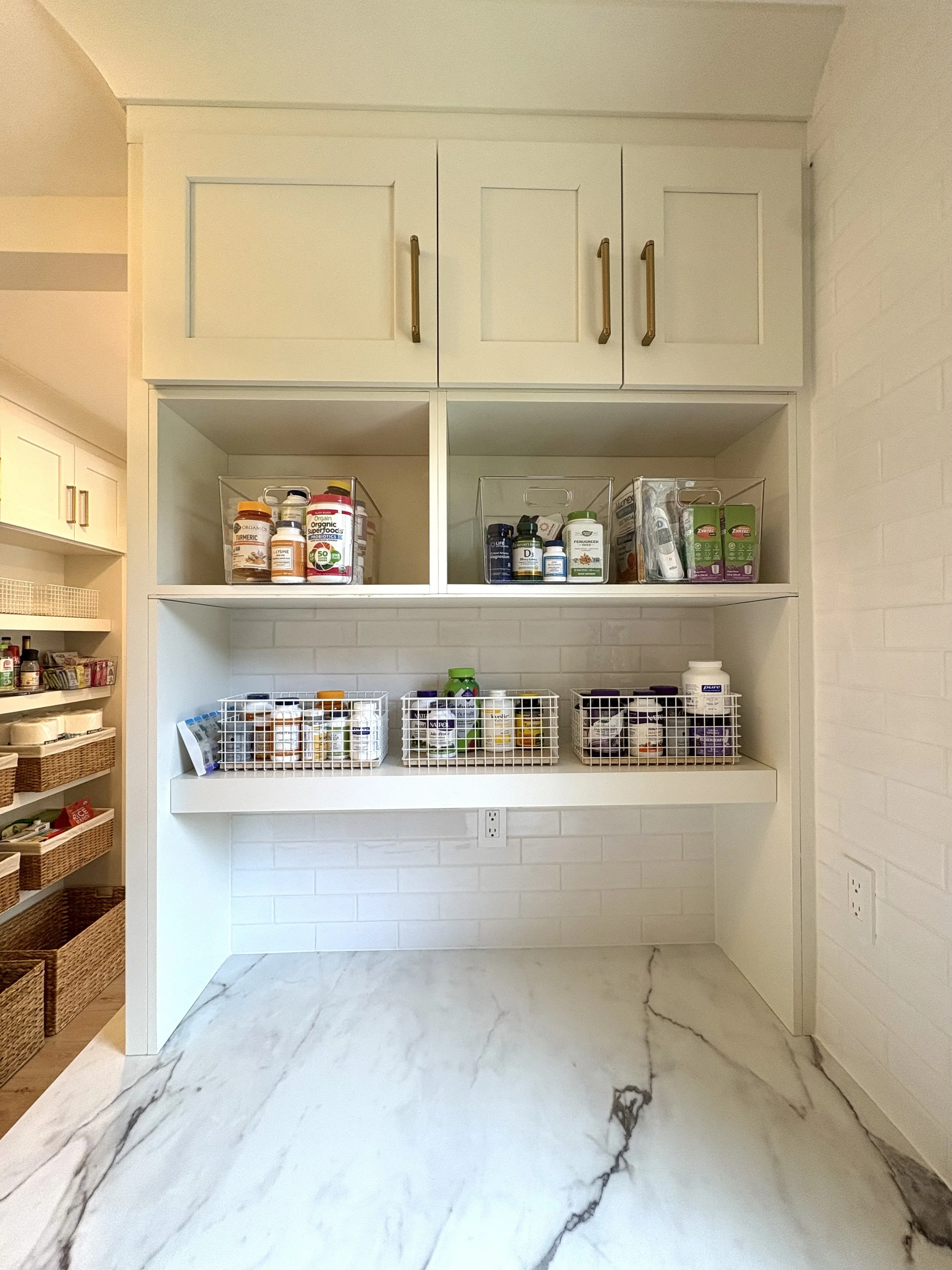 Kitchen cabinet with white open shelves containing bottles of dietary supplements, herbs, and vitamins, with a marble floor and white brick wall.