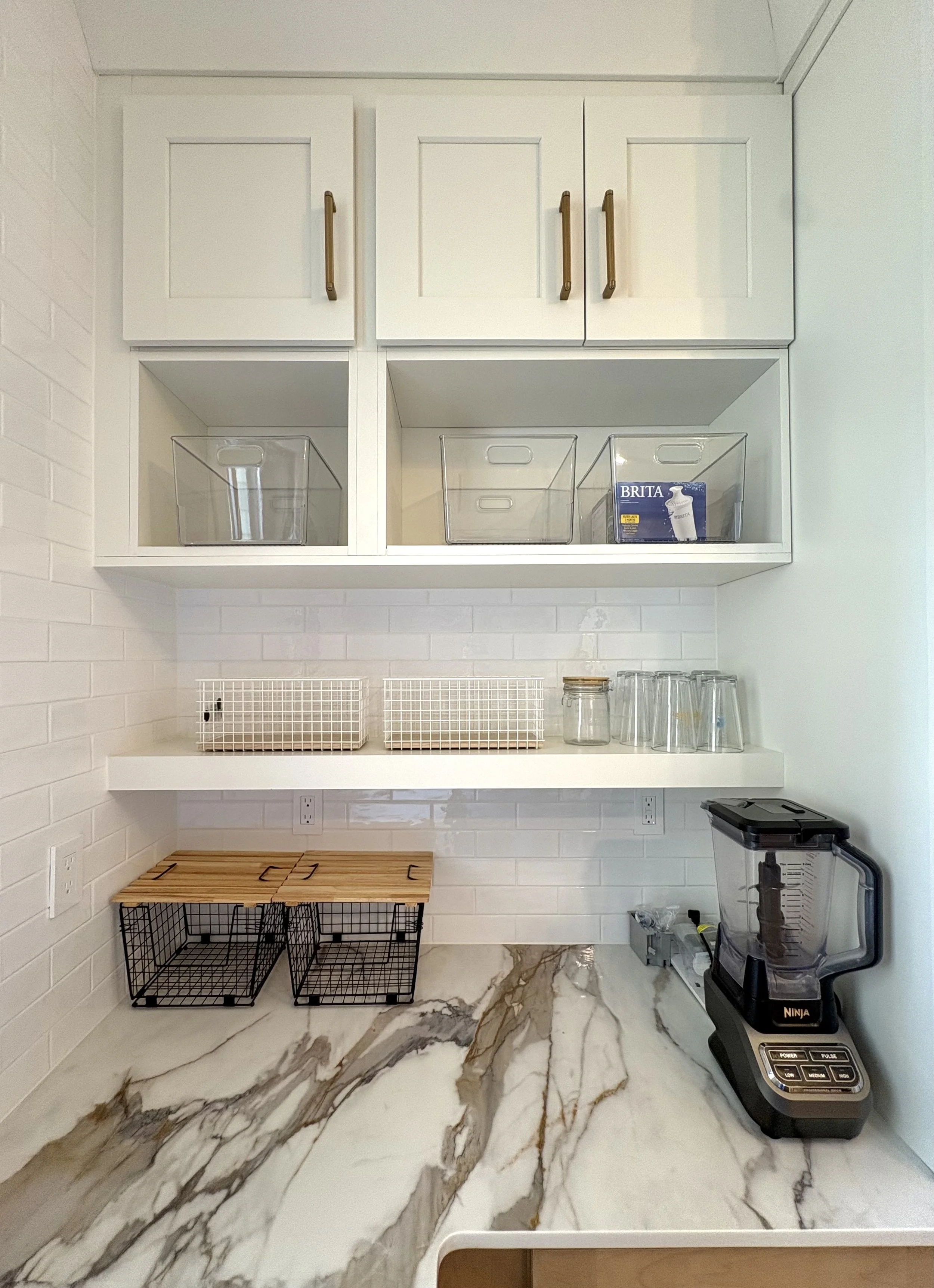 Kitchen counter with marble surface, black wire baskets, and a Ninja blender on the right side. Upper white cabinets with gold handles and open shelves with clear storage bins and a Brita water filter box. Wall with white brick tiles.