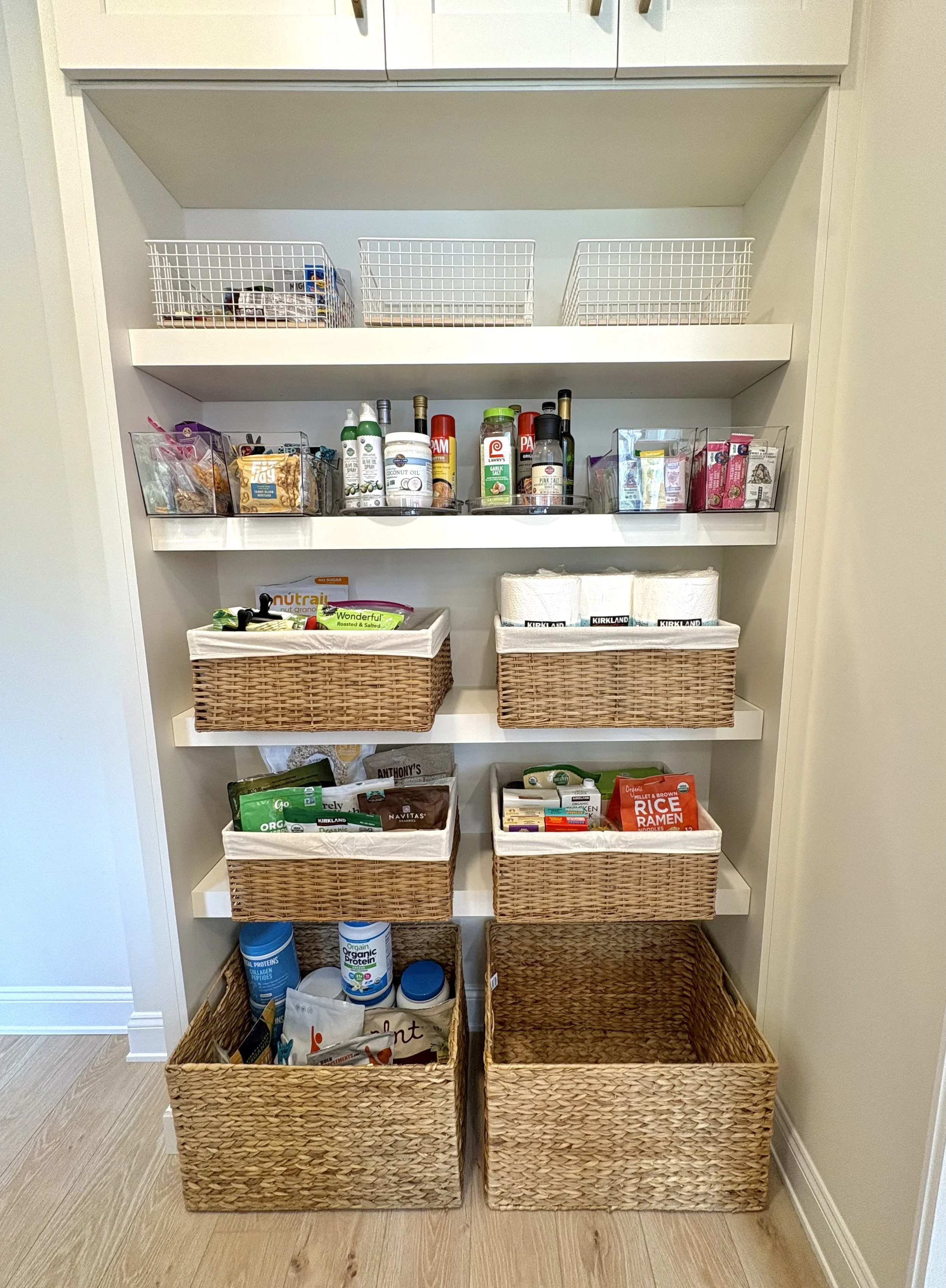 A pantry shelf with baskets and containers holding food, snacks, and household items.