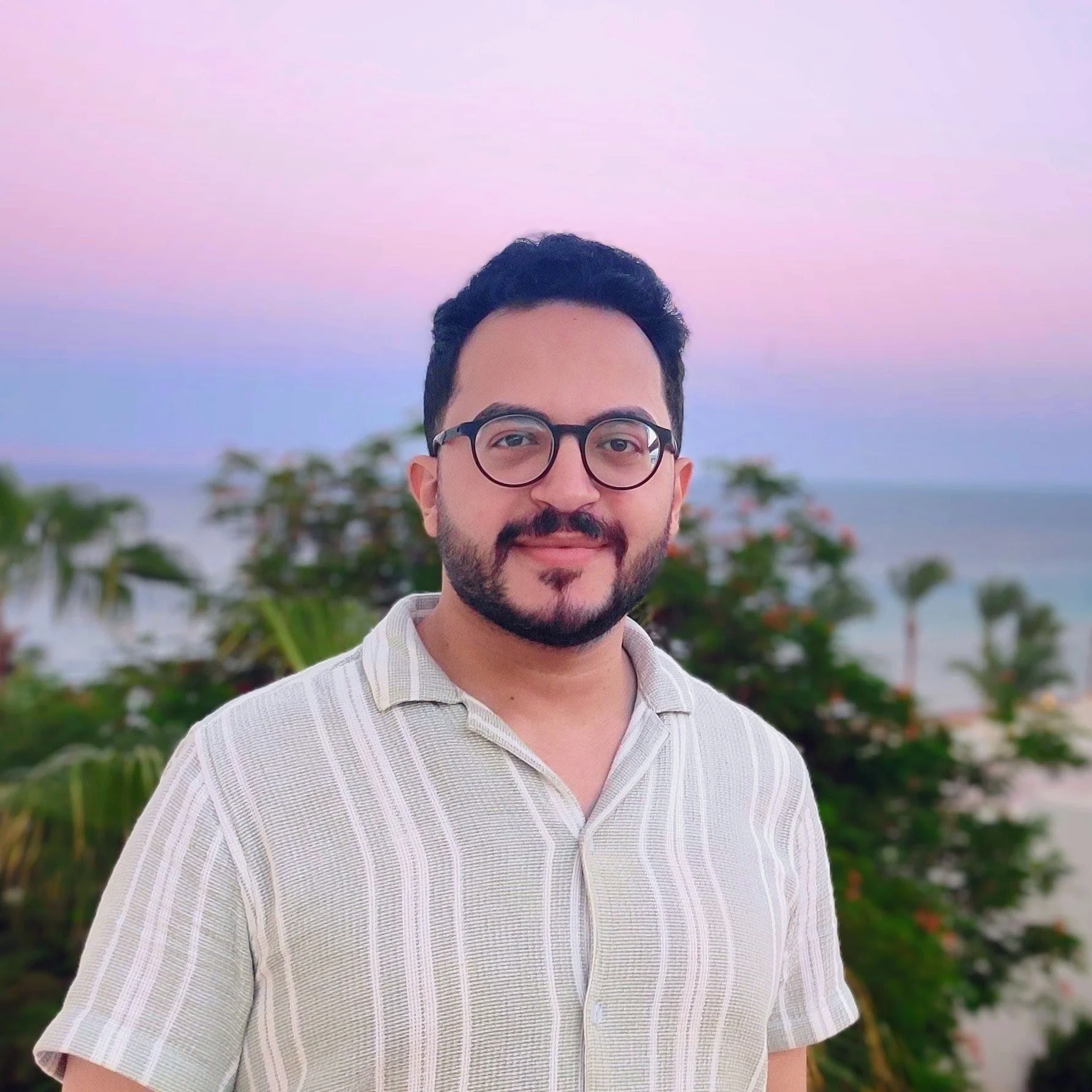 A man with glasses and facial hair smiling outdoors during sunset or dusk, with palm trees and the ocean in the background.