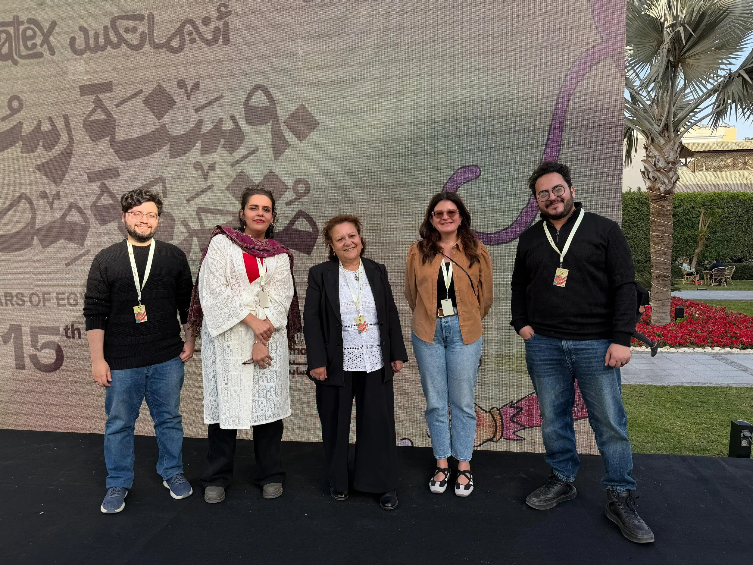 A group of five people standing outdoors in front of a large presentation board. Four women and one man, all smiling, wearing casual and semi-formal attire, with some wearing conference badges. There are palm trees and outdoor seating in the background.