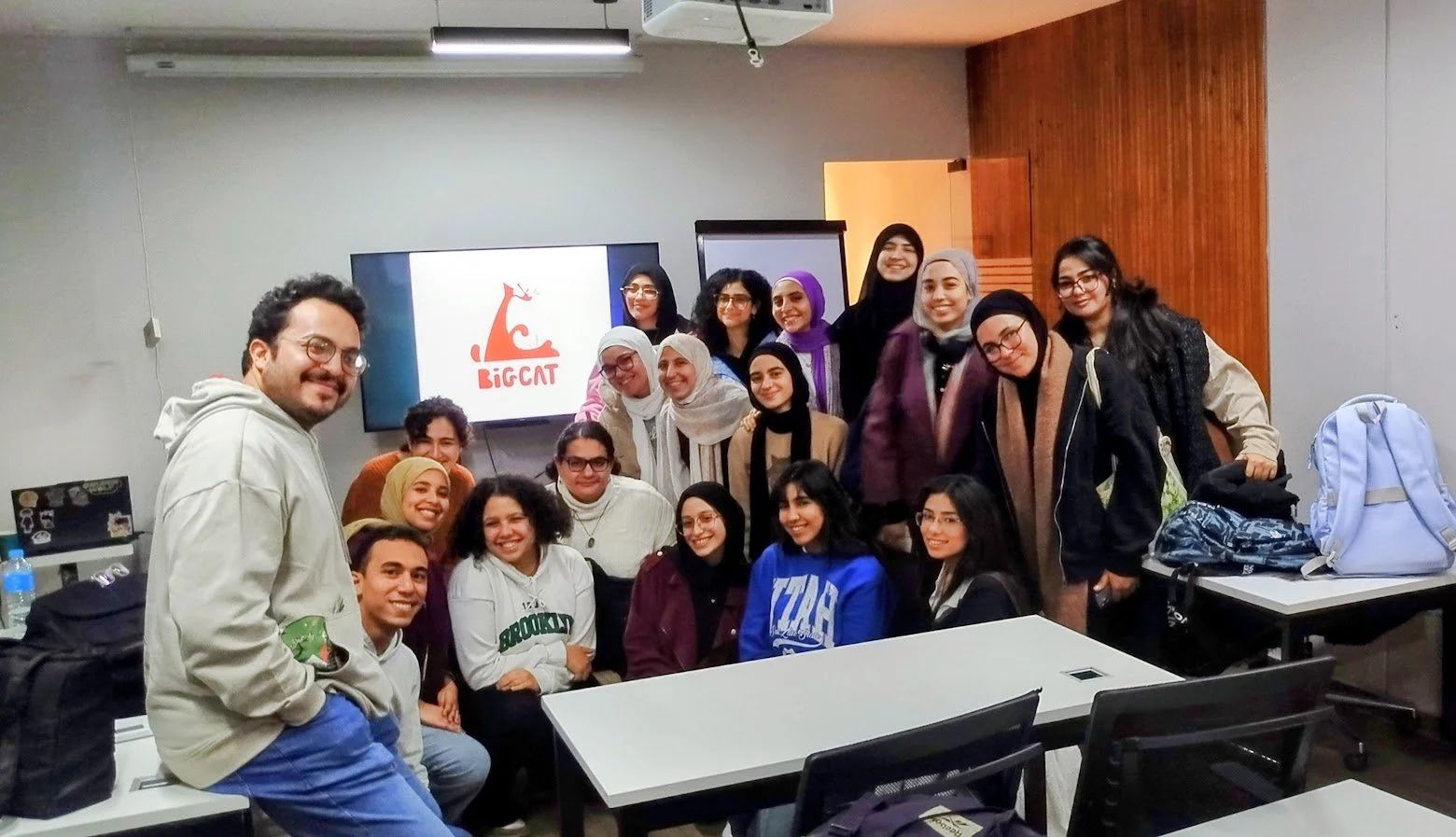 Group of diverse young women and one man smiling in a classroom or conference room, with a presentation screen displaying a red cartoon cat and the word "BIGCAT" in the background.