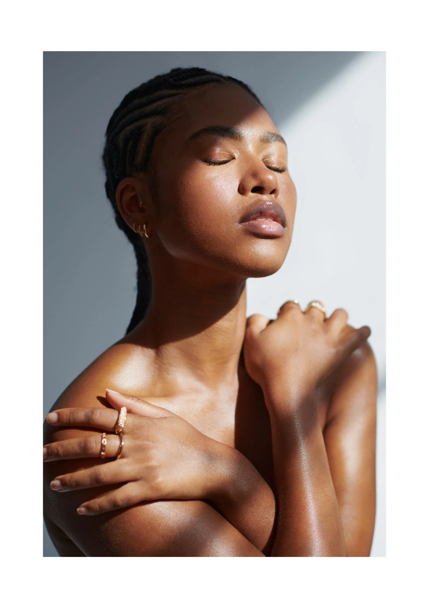 Close-up of a woman with eyes closed, embracing herself, with jewelry on her fingers, short braided hair, and smooth skin, in natural light.