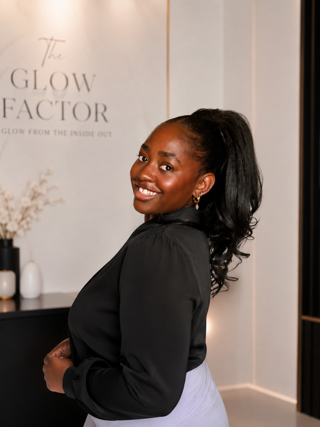 Smiling woman with dark hair tied back, wearing a black blouse and white pants, standing indoors near a sign that reads 'The Glow Factor' with decorative vases in the background.