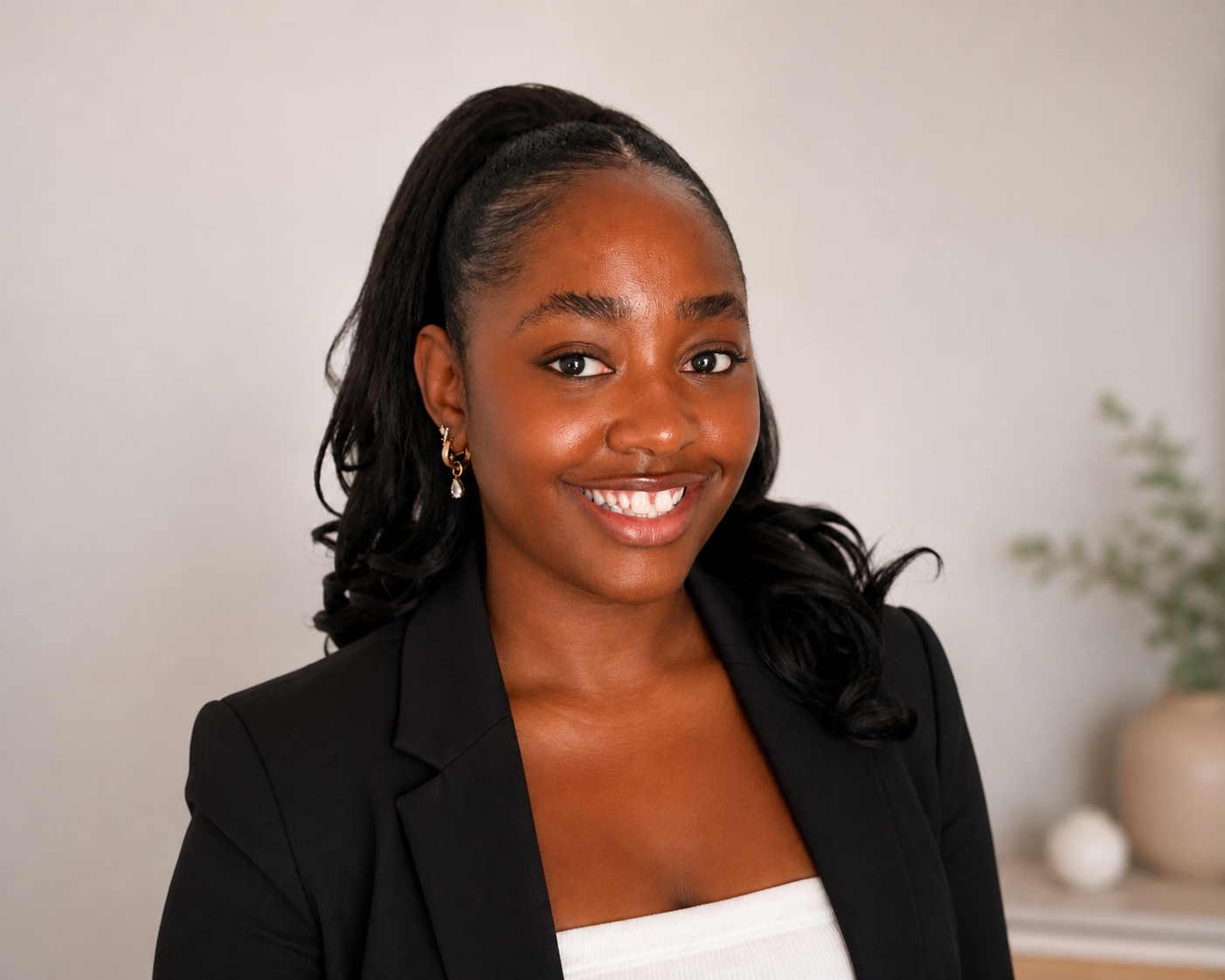 A smiling young African American woman with dark hair in loose curls, wearing a black blazer and white top, standing in a room with neutral decor and a blurred plant in the background.