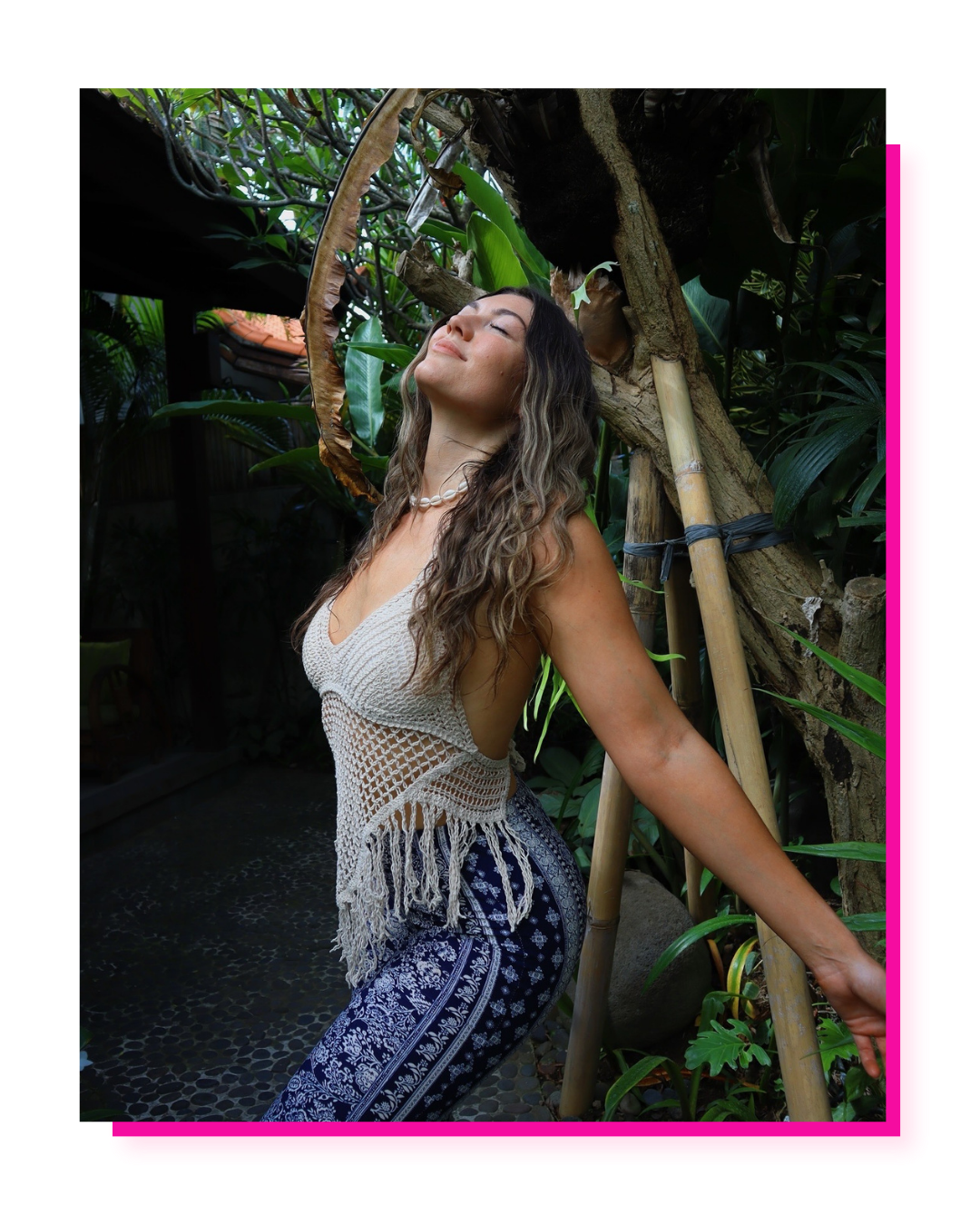 Woman with long, wavy hair wearing a crochet top and patterned pants, smiling with eyes closed, in a lush garden setting with tropical plants and bamboo.