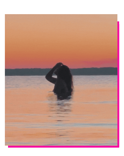 A person standing in water during sunset, with one hand running through their hair.