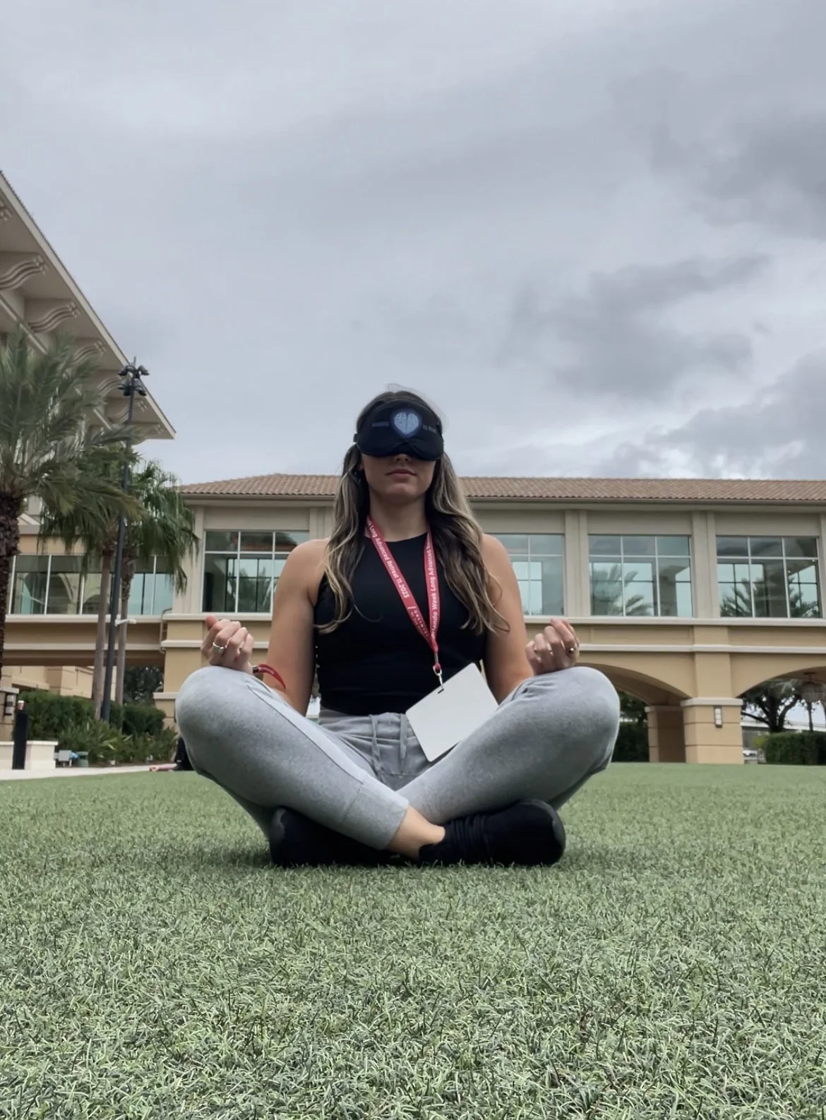 Woman sitting cross-legged on grass, practicing meditation or yoga outdoors, wearing leggings, tank top, an eye mask, and a lanyard with an ID badge, with a building and cloudy sky in the background.