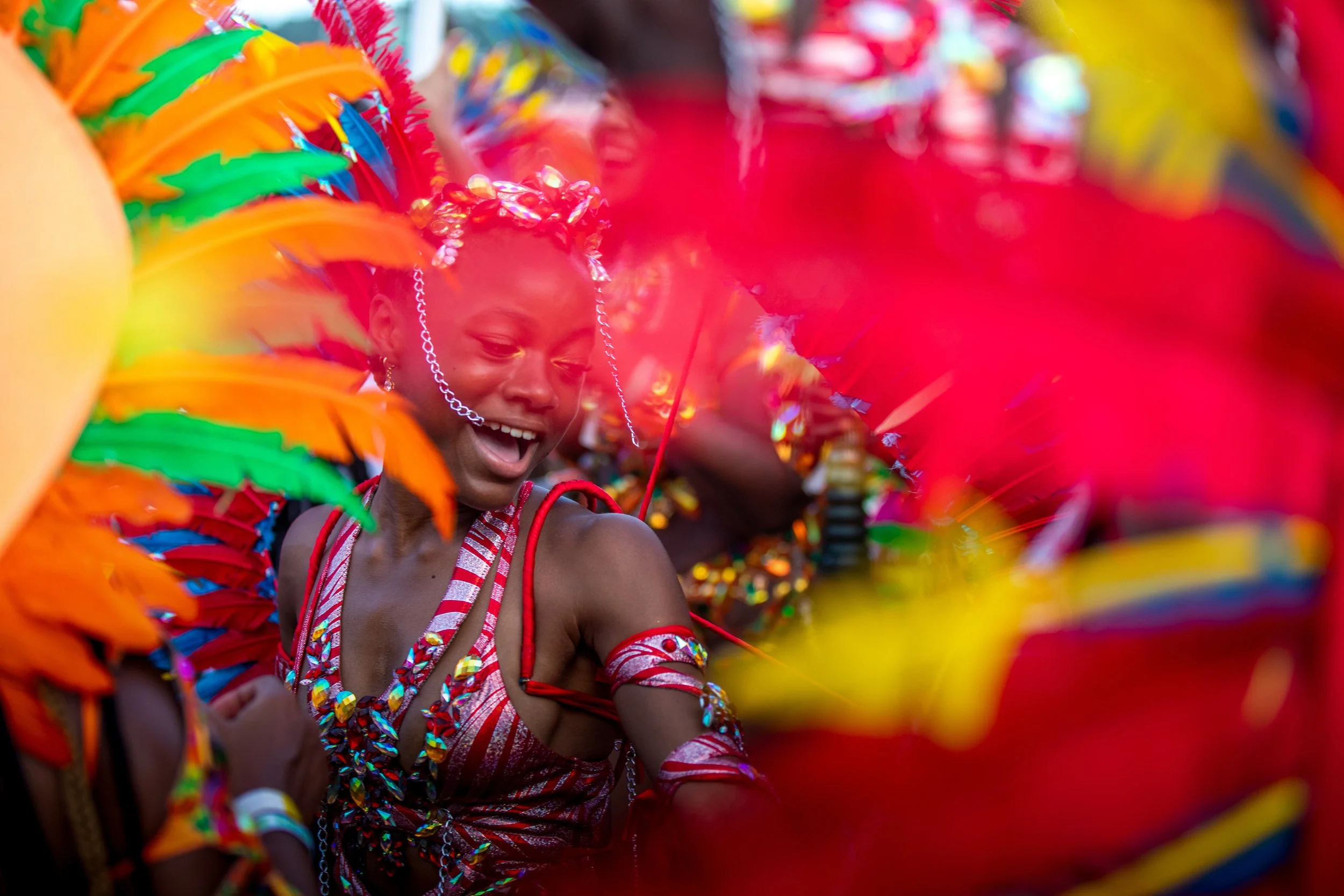 A young person wearing colorful, sequined Mardi Gras costume and a crown is dancing and smiling at a lively parade.
