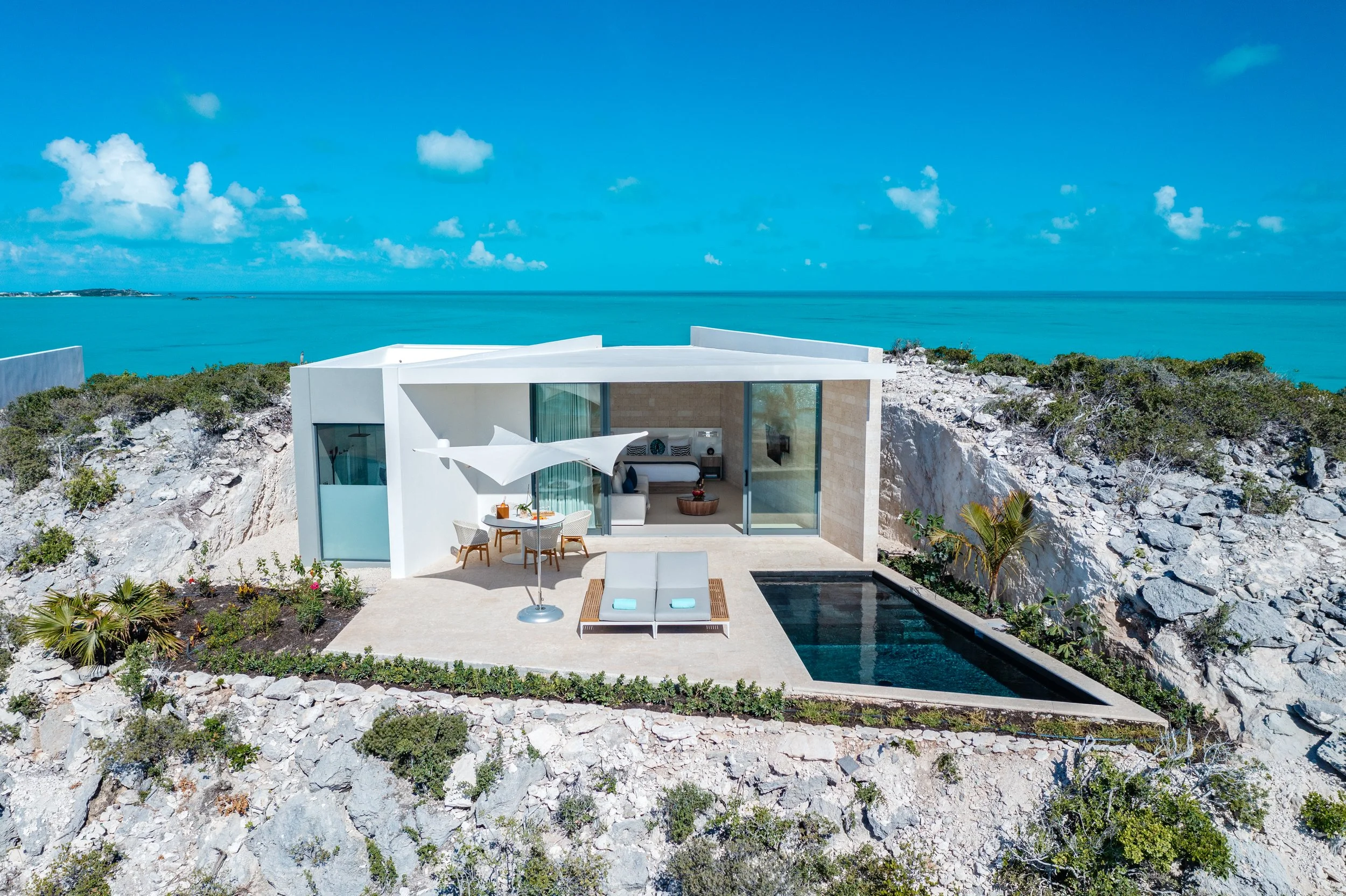 Modern white house with outdoor seating area, built into a rocky hillside overlooking the ocean with clear blue sky.