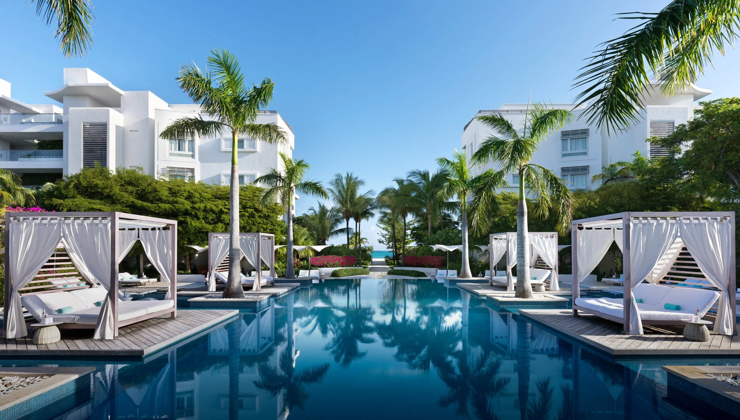 Luxury resort pool area with palm trees, white cabanas, and modern white buildings under a clear blue sky.