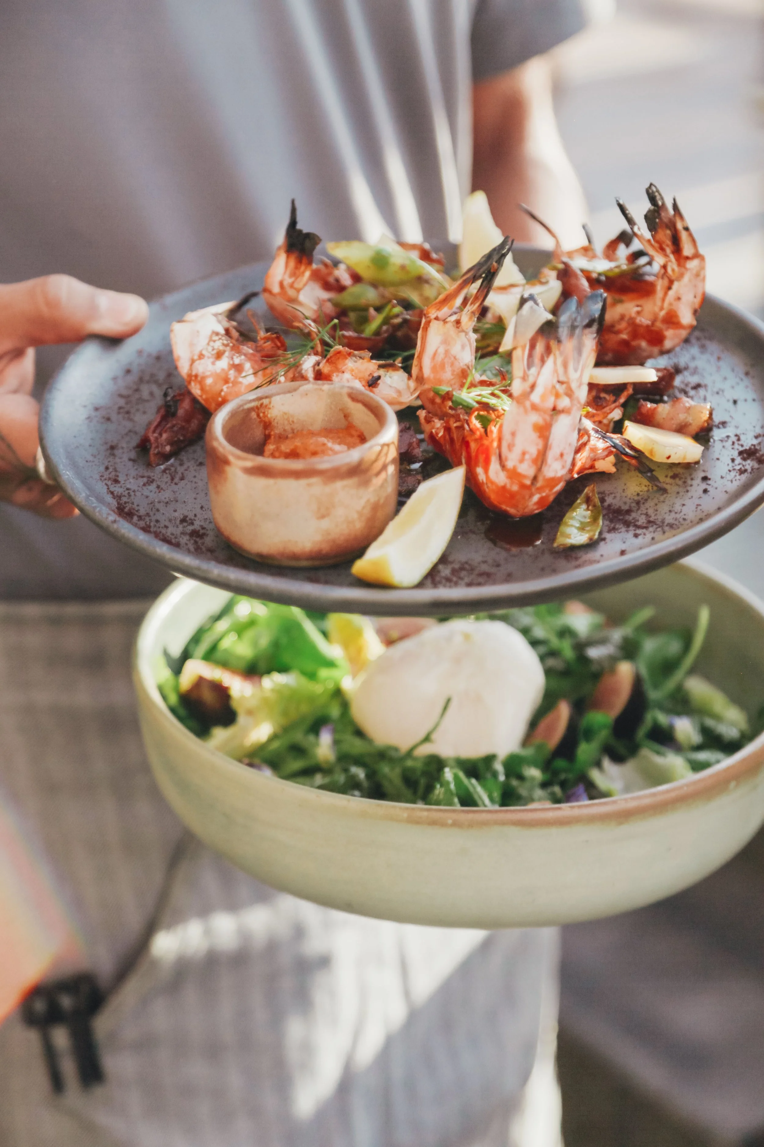 A person holding a plate with cooked grilled shrimp garnished with herbs and lemon wedges, with a bowl of salad below.