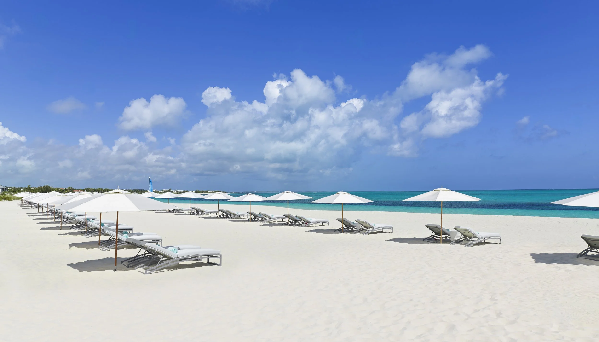 Empty white sandy beach with white umbrellas and lounge chairs facing the turquoise ocean under a blue sky with scattered white clouds.
