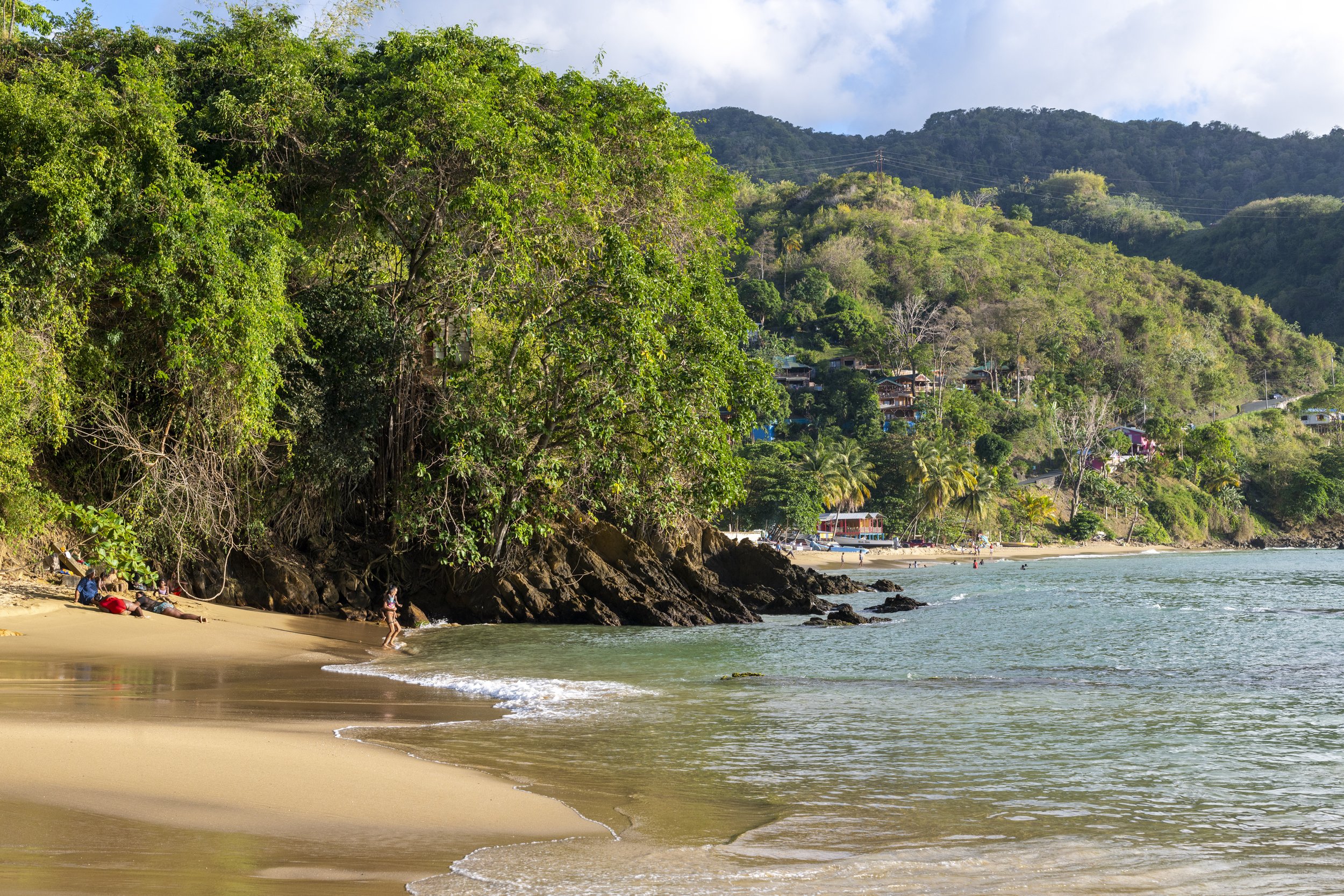 A tropical beach with golden sand, turquoise water, and lush green hills in the background. There are people relaxing on the beach and swimming in the water, with a few houses visible on the hillside.
