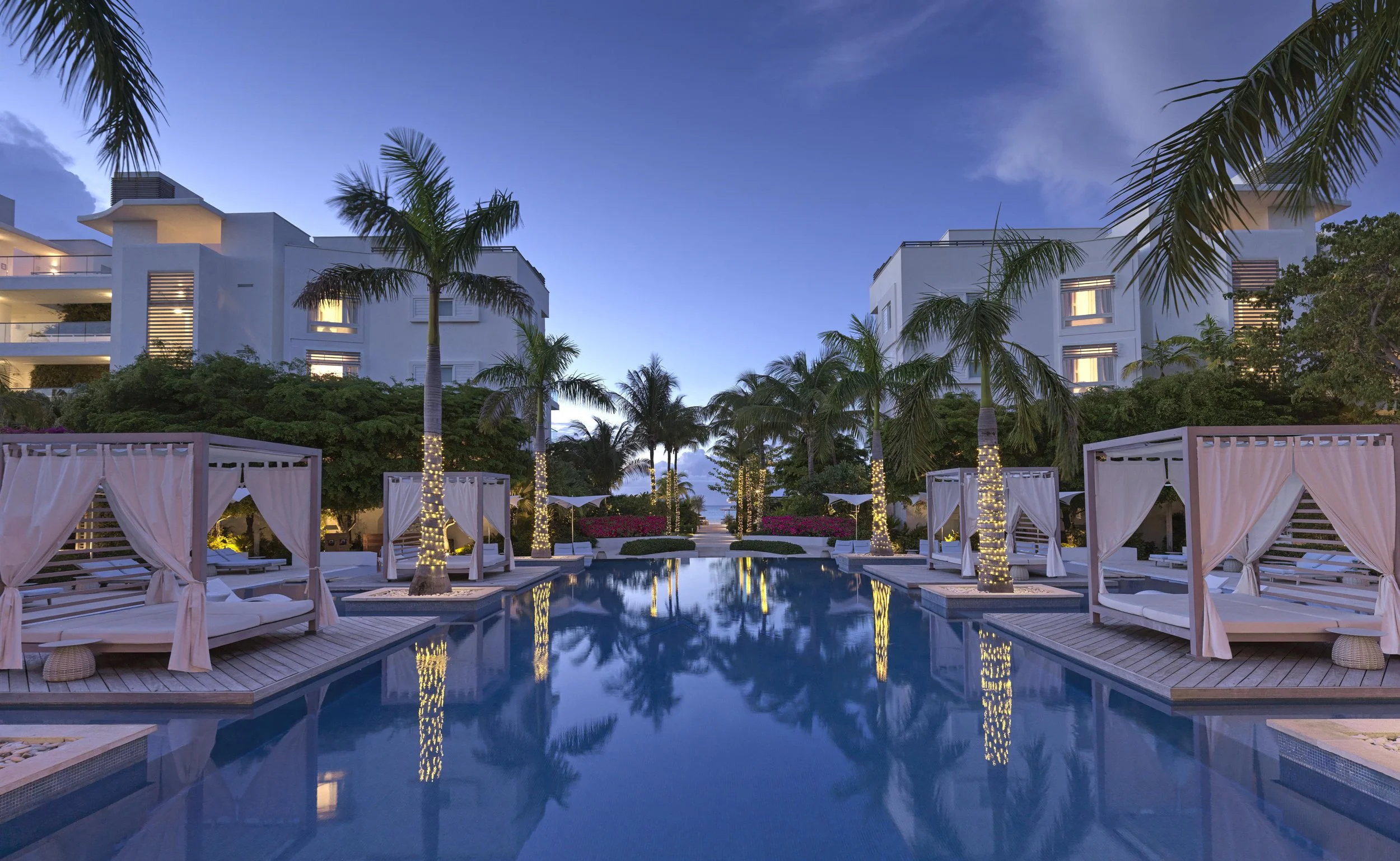 A luxurious outdoor pool area at dusk with palm trees decorated with string lights, white cabanas with curtains, and modern white residential buildings in the background under a partly cloudy sky.
