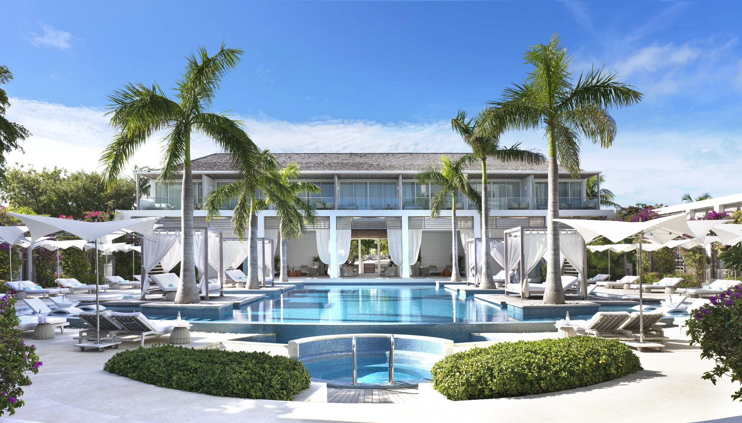 Luxury resort with a swimming pool, surrounded by palm trees, lounge chairs, and cabanas under umbrellas, with a modern villa in the background set against a bright blue sky.