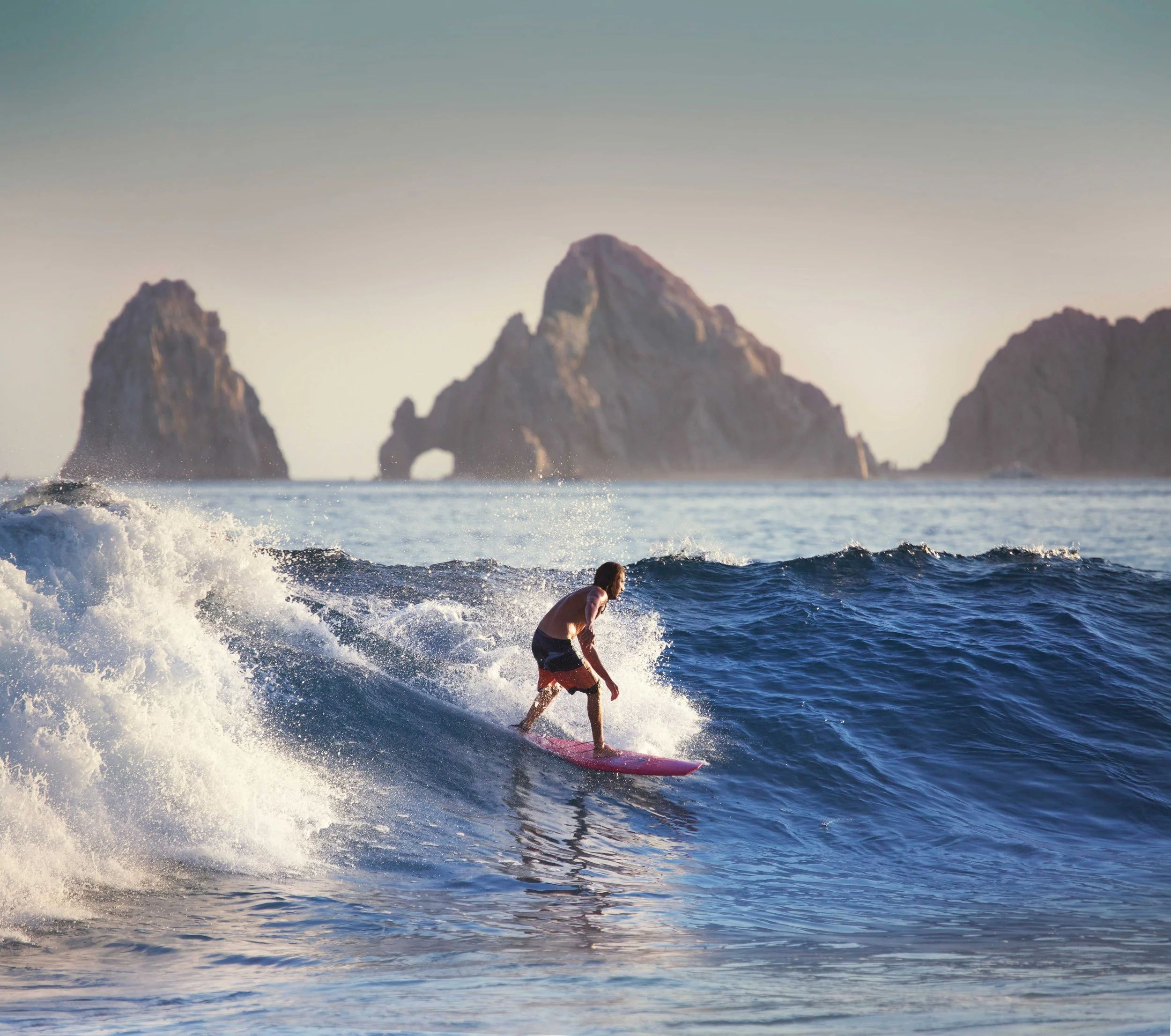A person surfing on a pink surfboard in the ocean near rocky islands, with waves splashing and a clear sky.