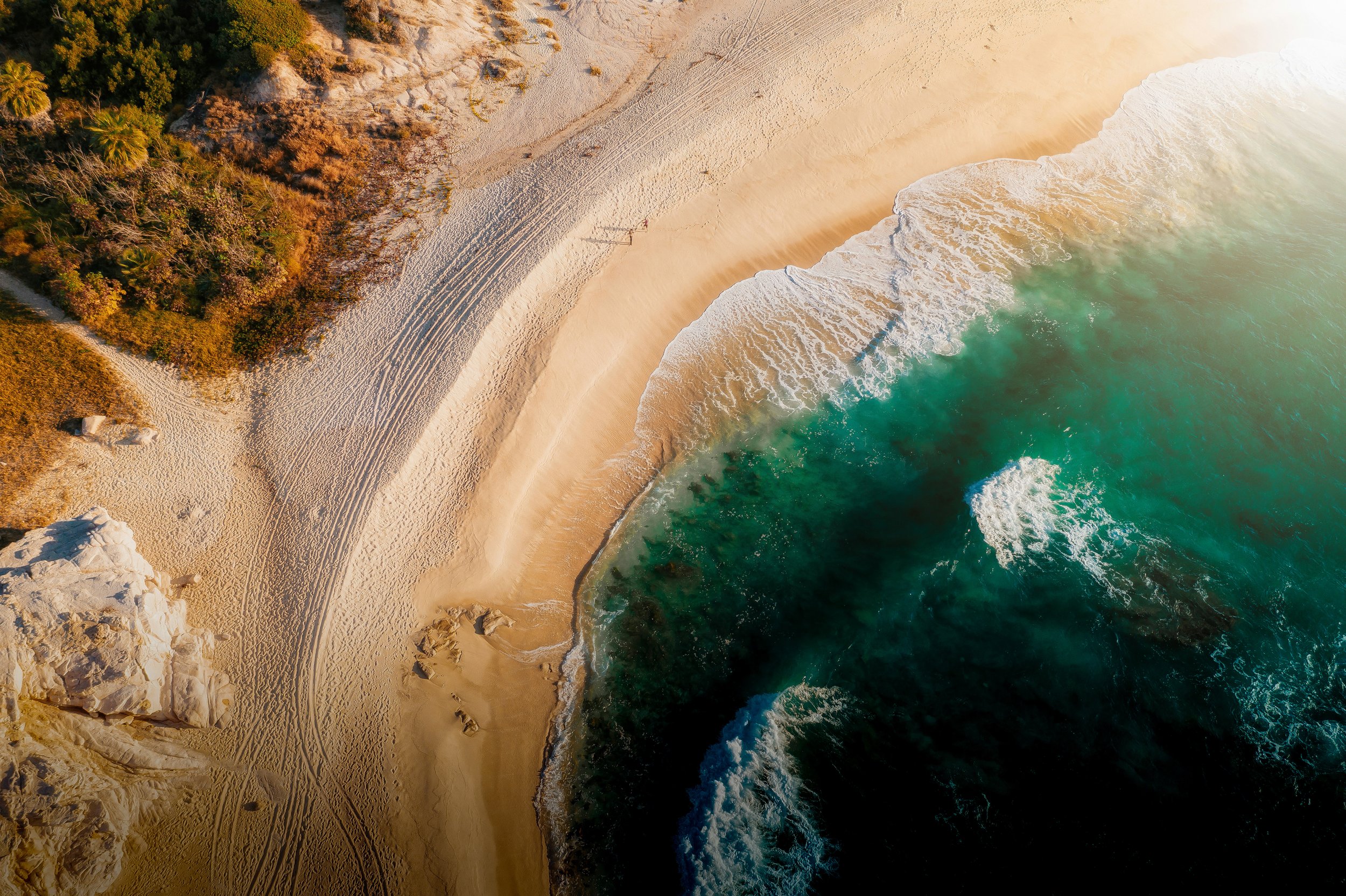 An aerial view of a sandy beach with footprints and tire tracks, backed by a lush green forest on the left, and turquoise ocean waves crashing onto the shore on the right.