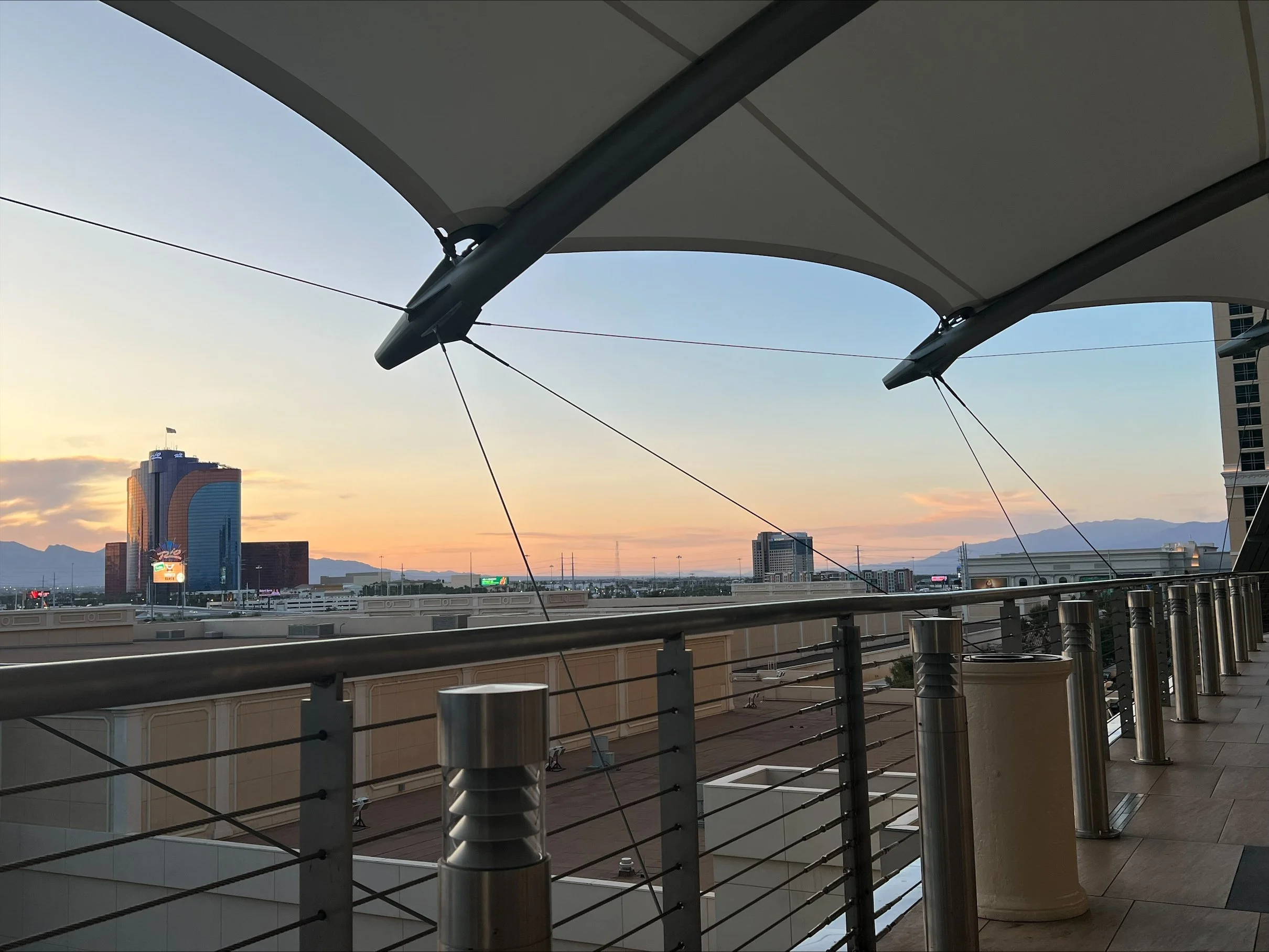 View from an outdoor balcony or rooftop with metal railings, large light-colored columns, and a partial ceiling structure. In the background, there are high-rise buildings, mountains, and a sunset sky with soft clouds and a warm glow.
