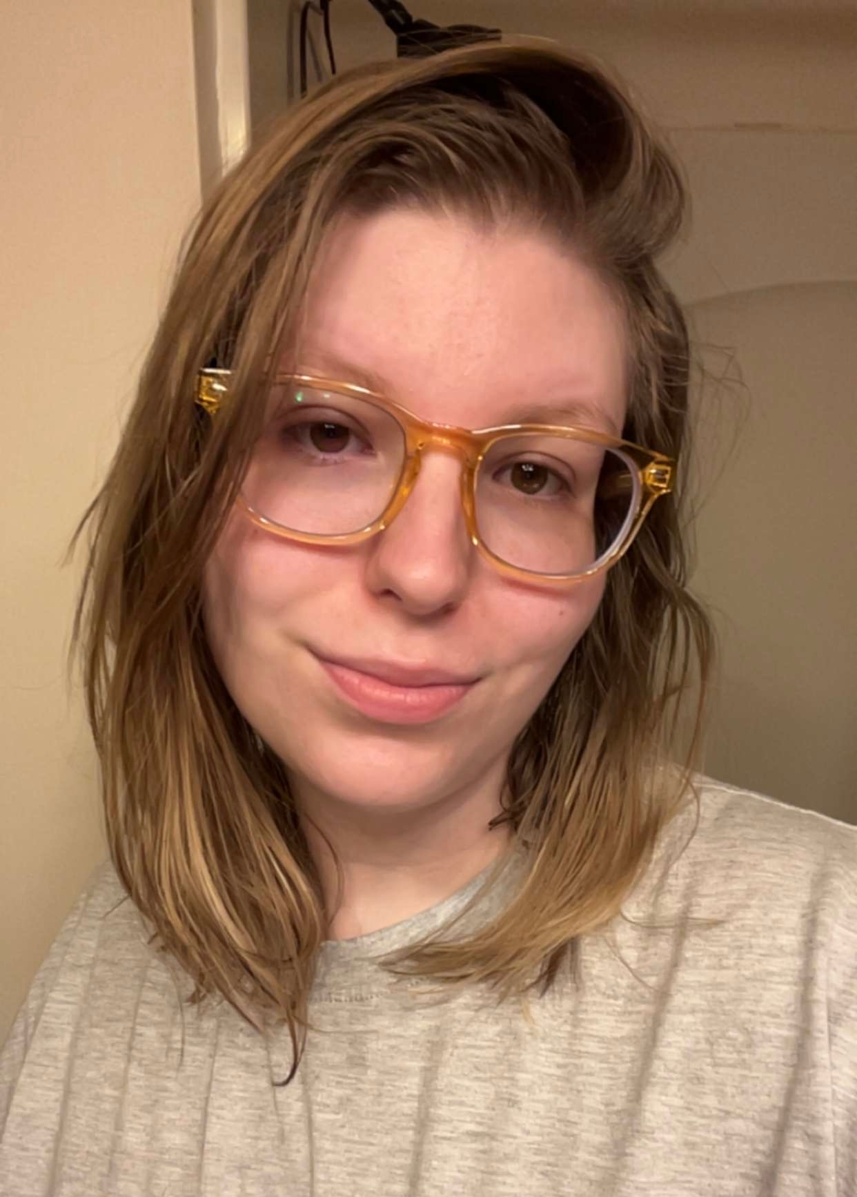 A young woman with shoulder-length light brown hair, wearing large, amber-colored glasses, and a light gray shirt, smiling gently in an indoor setting.