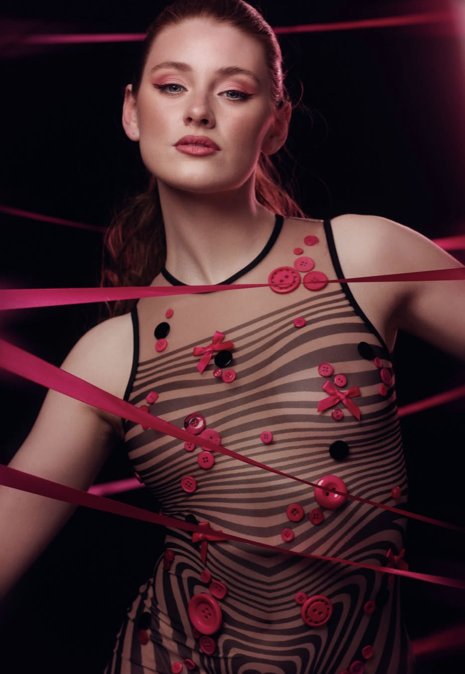 A woman with red hair styled in a ponytail, wearing a sheer dress decorated with pink and black buttons and ribbons, posing against a dark background with pink light streaks.