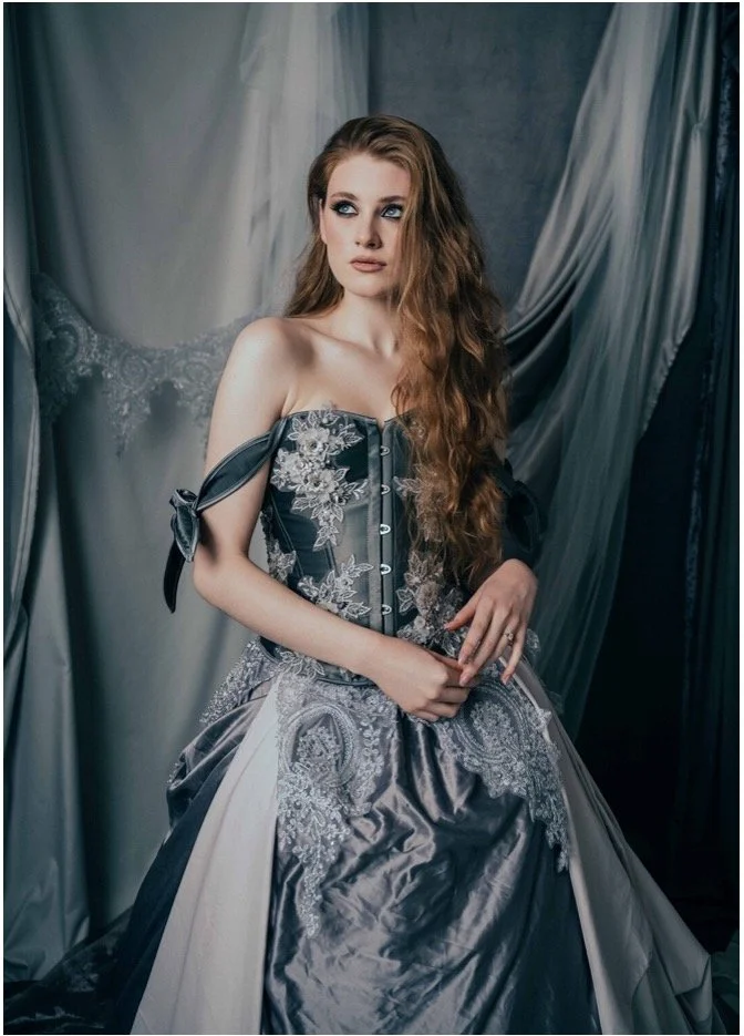 A young woman with long, curly auburn hair is wearing an elegant, off-the-shoulder gray gown with lace and embroidery details. She is posed in front of a draped, fabric backdrop, gazing to the side with a thoughtful expression.