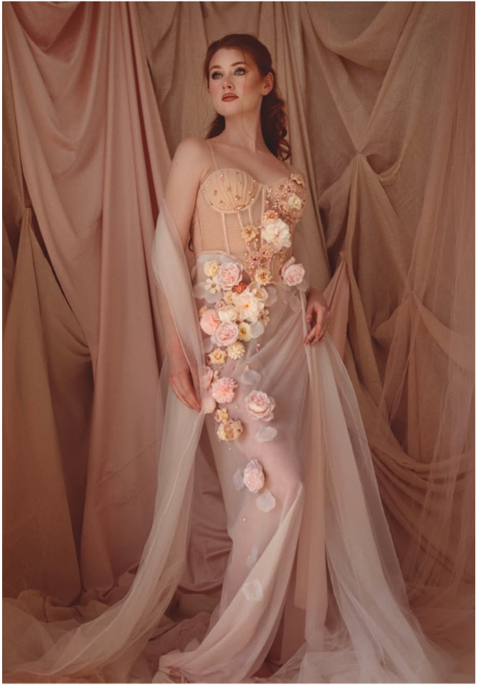 A woman in a flowing, nude-colored gown adorned with flowers and tulle, standing against a backdrop of pinkish-beige draped fabric.