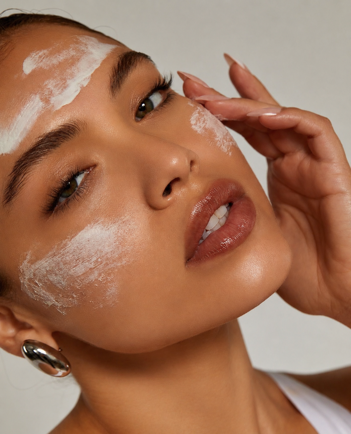 Close-up of a woman with white cream on her face, gently touching her face with her hand, with glossy lips and hoop earrings.