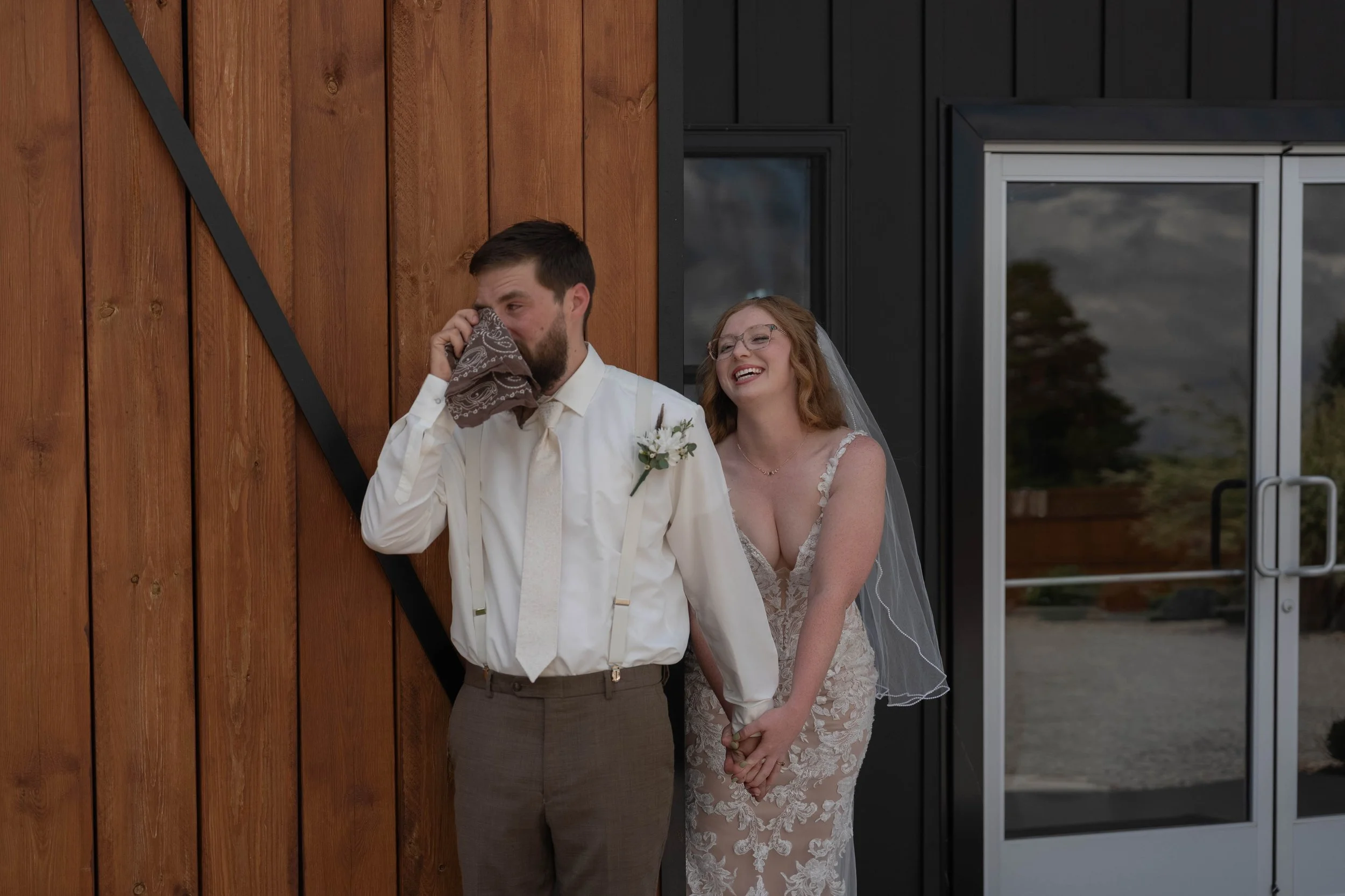 A newlywed couple holding hands, with the groom sneezing into a tissue and the bride smiling behind him, standing in front of a wooden wall and a glass door.