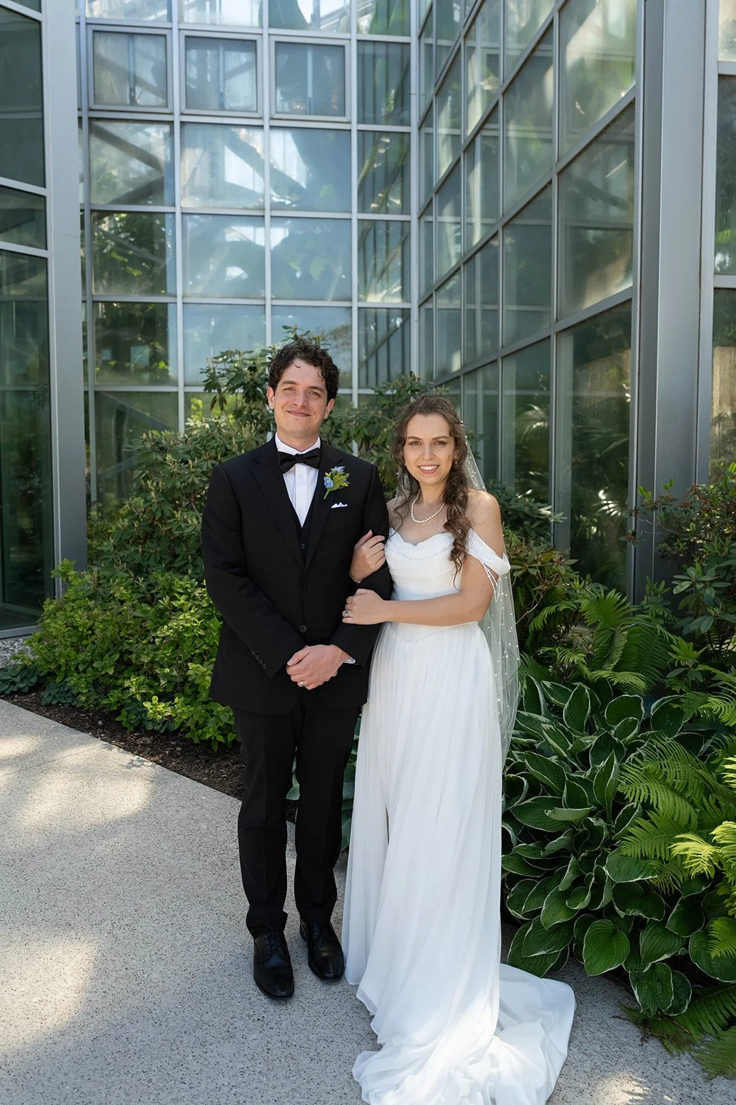 A newlywed couple stands outdoors in front of a glass greenhouse, with lush green plants and shrubs, dressed in wedding attire. The groom wears a black tuxedo with a bow tie, and the bride wears a white gown with a veil and pearl necklace.