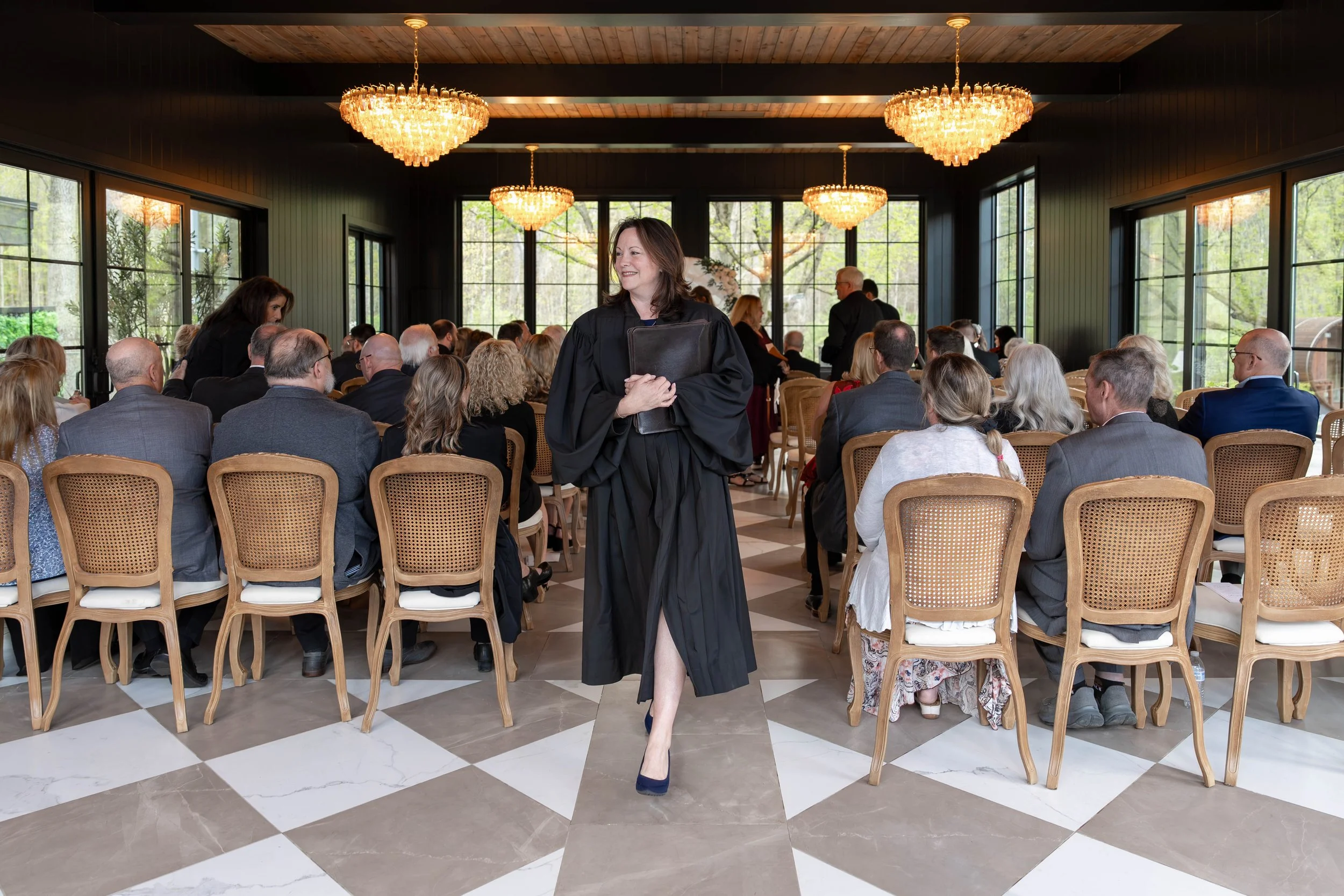 A woman in a black gown and heels walking in a room filled with seated attendees at a formal event, with large windows and chandeliers overhead.