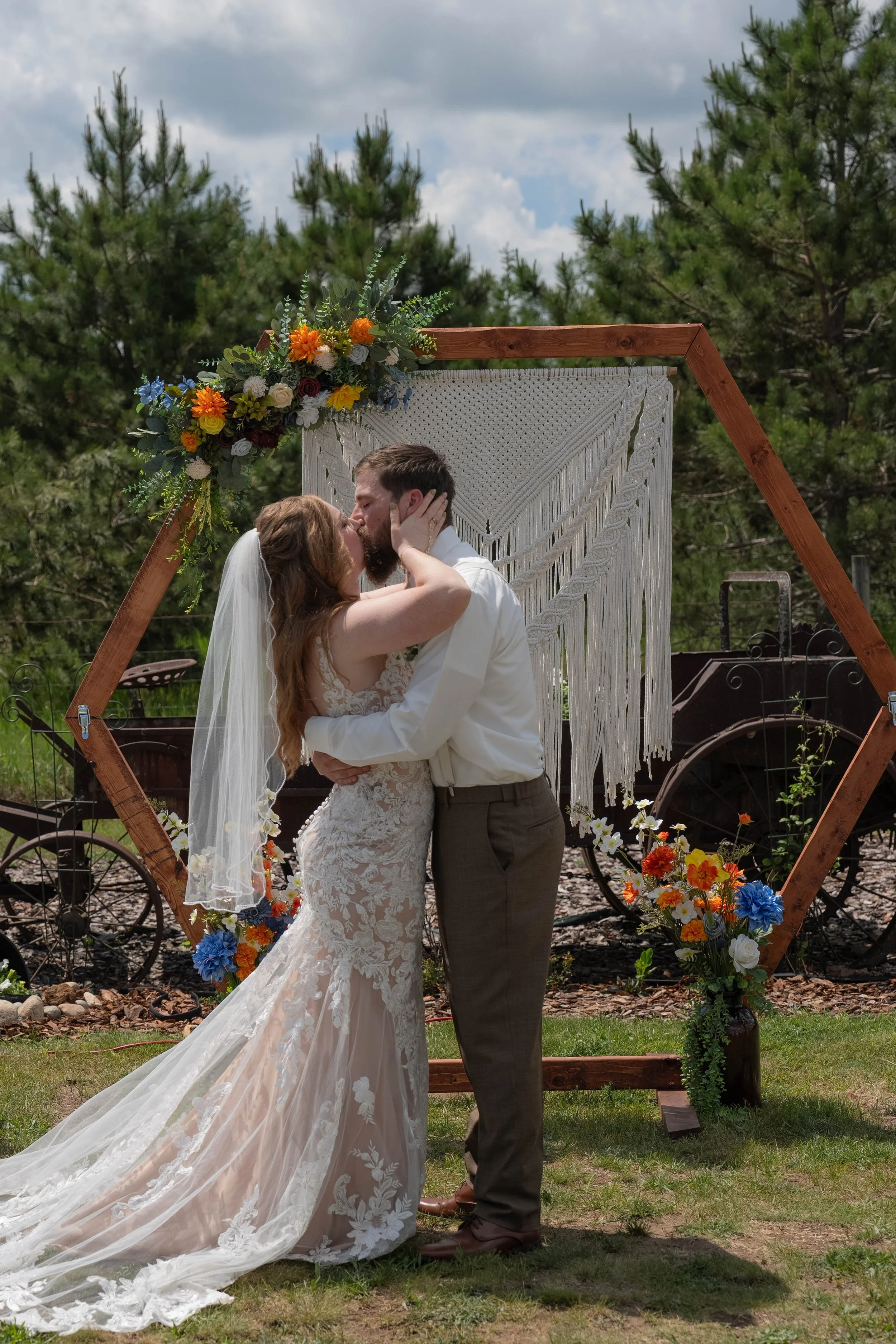 A bride and groom sharing a kiss at their outdoor wedding. The bride wears a lace wedding gown and veil, and the groom is in a gray suit. They stand in front of a rustic wooden frame decorated with colorful flowers and greenery, with trees and a clou