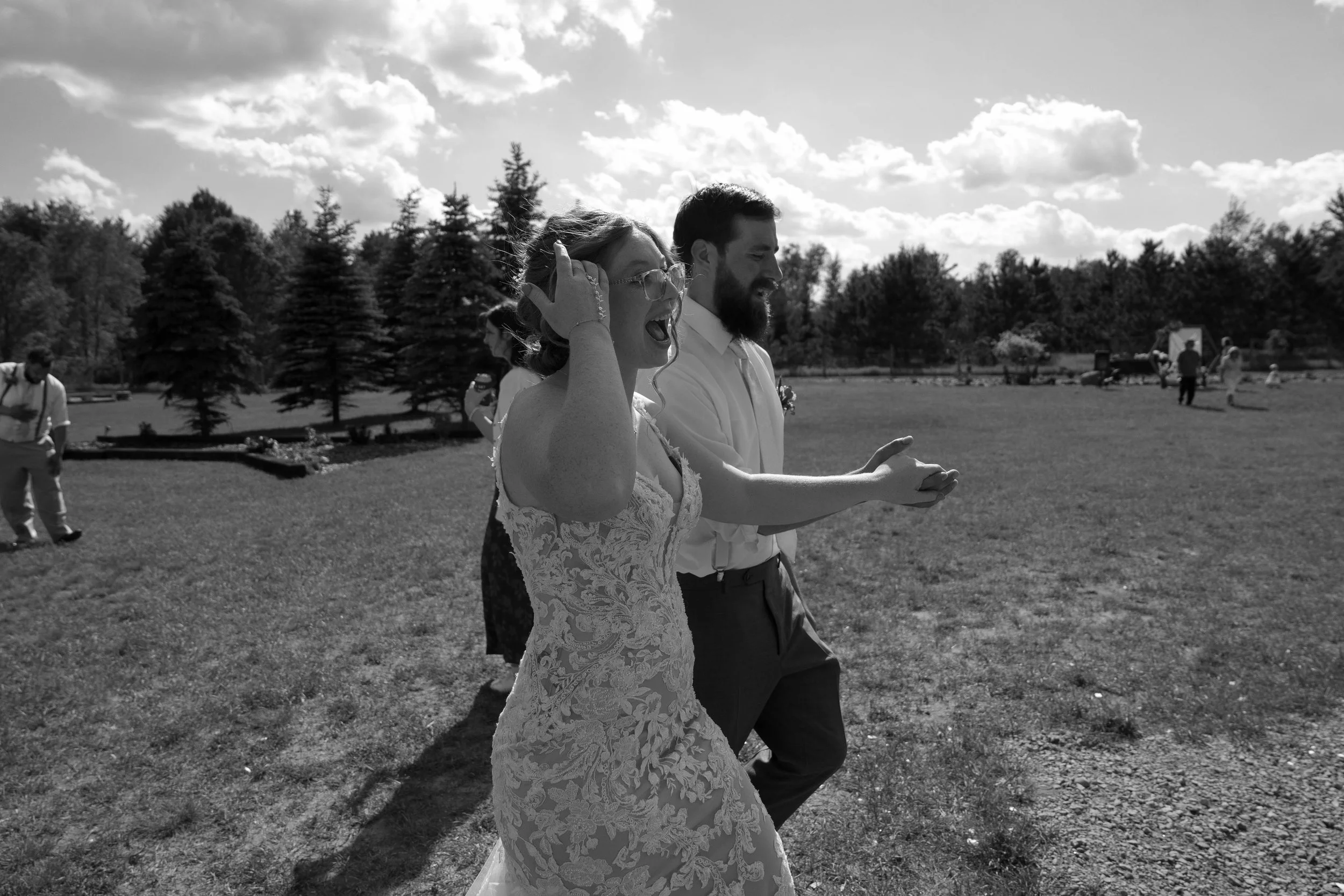 Black and white photo of a wedding couple holding hands and walking outdoors on a sunny day with trees and clouds in the background.