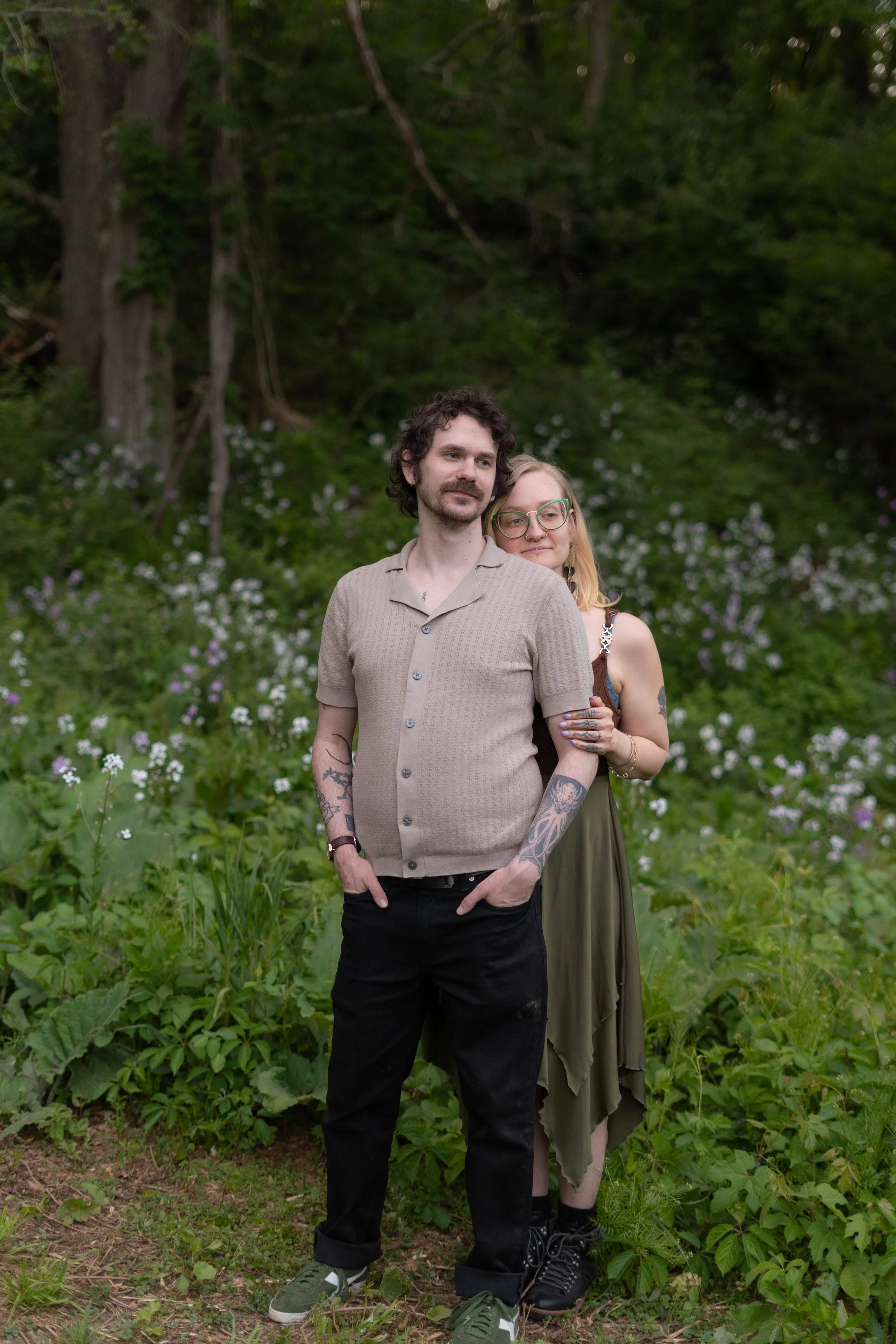 A man with dark, curly hair and tattooed arms standing in front of a woman with blonde hair, glasses, and tattoos, in a green, floral outdoor setting.
