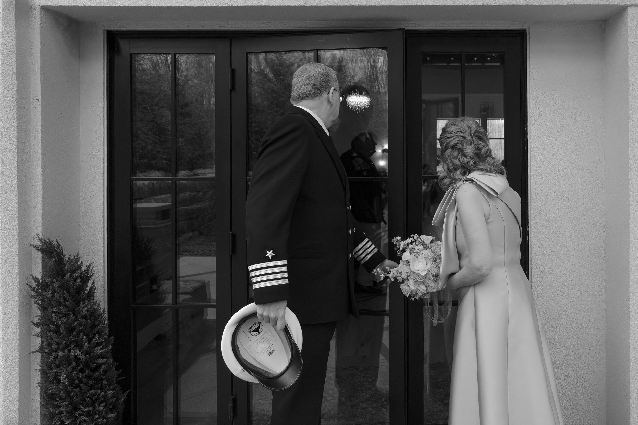 A man in a military uniform holding a hat and a woman in a white dress holding a bouquet of flowers standing on a porch in front of glass doors.