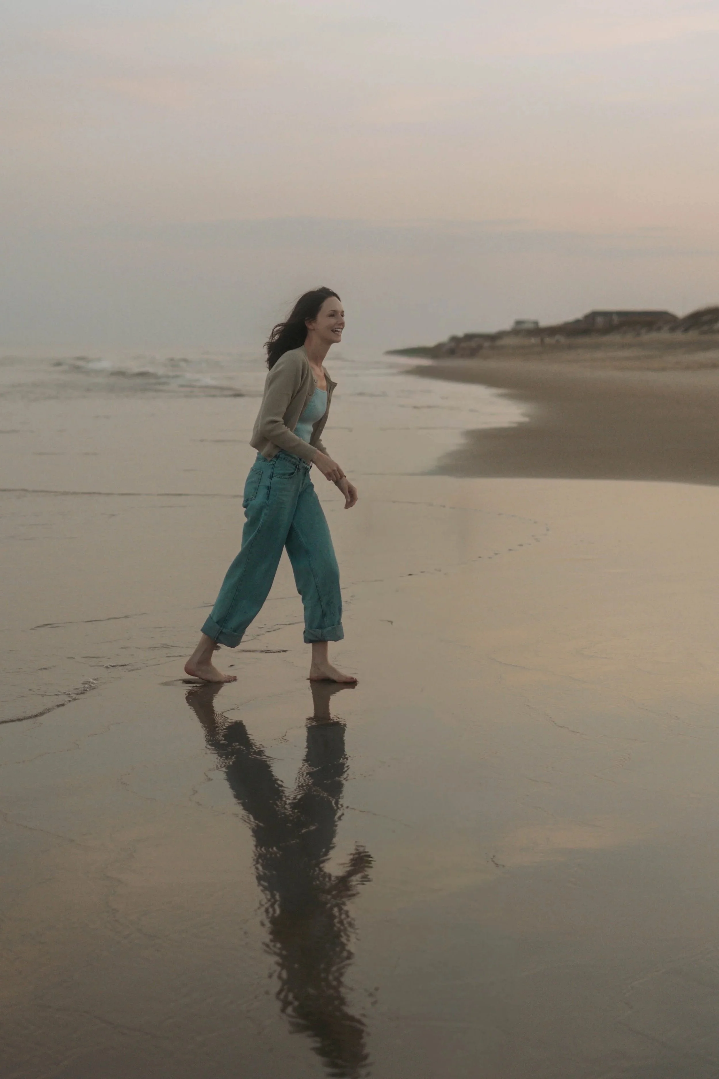 A young woman with dark hair smiling and walking barefoot on the wet sand of a beach during sunset, with her reflection visible on the shallow water, and a distant shoreline with houses in the background.