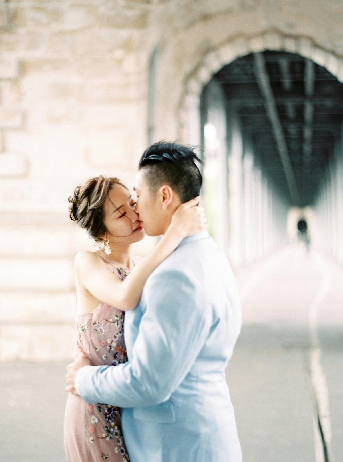 A couple kissing under a bridge, with the woman in a floral dress and the man in a light suit, in an urban setting.
